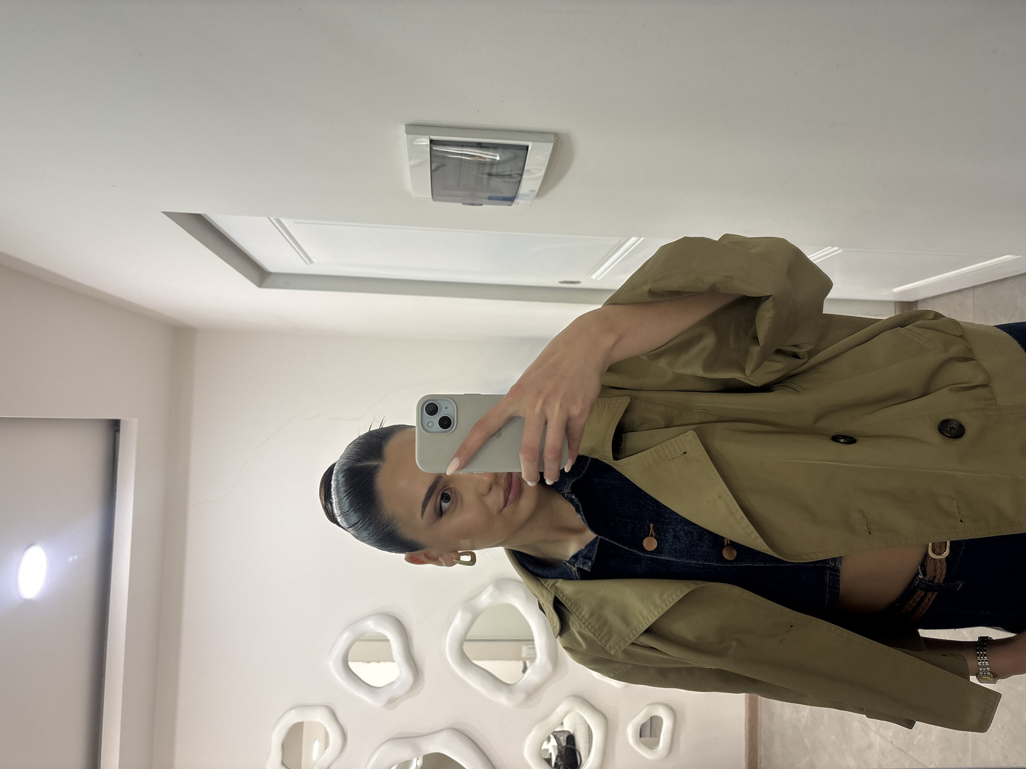 A woman is taking a selfie in what appears to be a restroom. She is wearing a khaki trench coat over a denim shirt and black pants with a brown belt. Her hair is pulled back in a bun, and she has one gold earring visible. She is holding a smartphone to take the picture.
The background features a white wall decorated with several organically shaped white mirrors. There is also a white door and a white ceiling with a rectangular vent.
While the location in Yerevan, Armenia, is mentioned, there are no discernible elements in the image to directly verify that specific geographical location. Therefore, only the visible information from the image can be described factually.