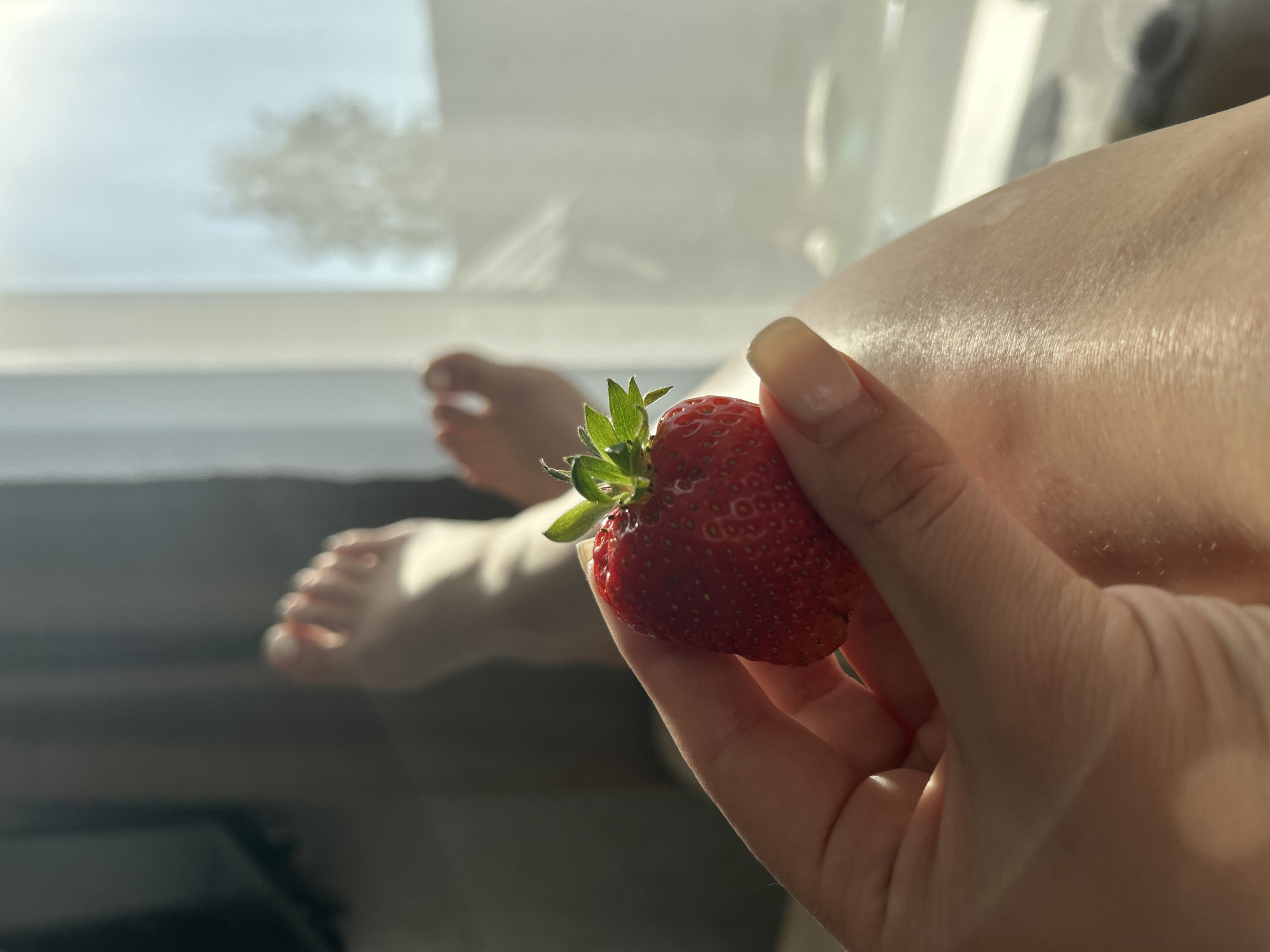 A hand with manicured nails holds a strawberry in the foreground. In the background, a window provides a blurred view, with feet and legs visible beside the window. The location is Chaikovsky, Russia.