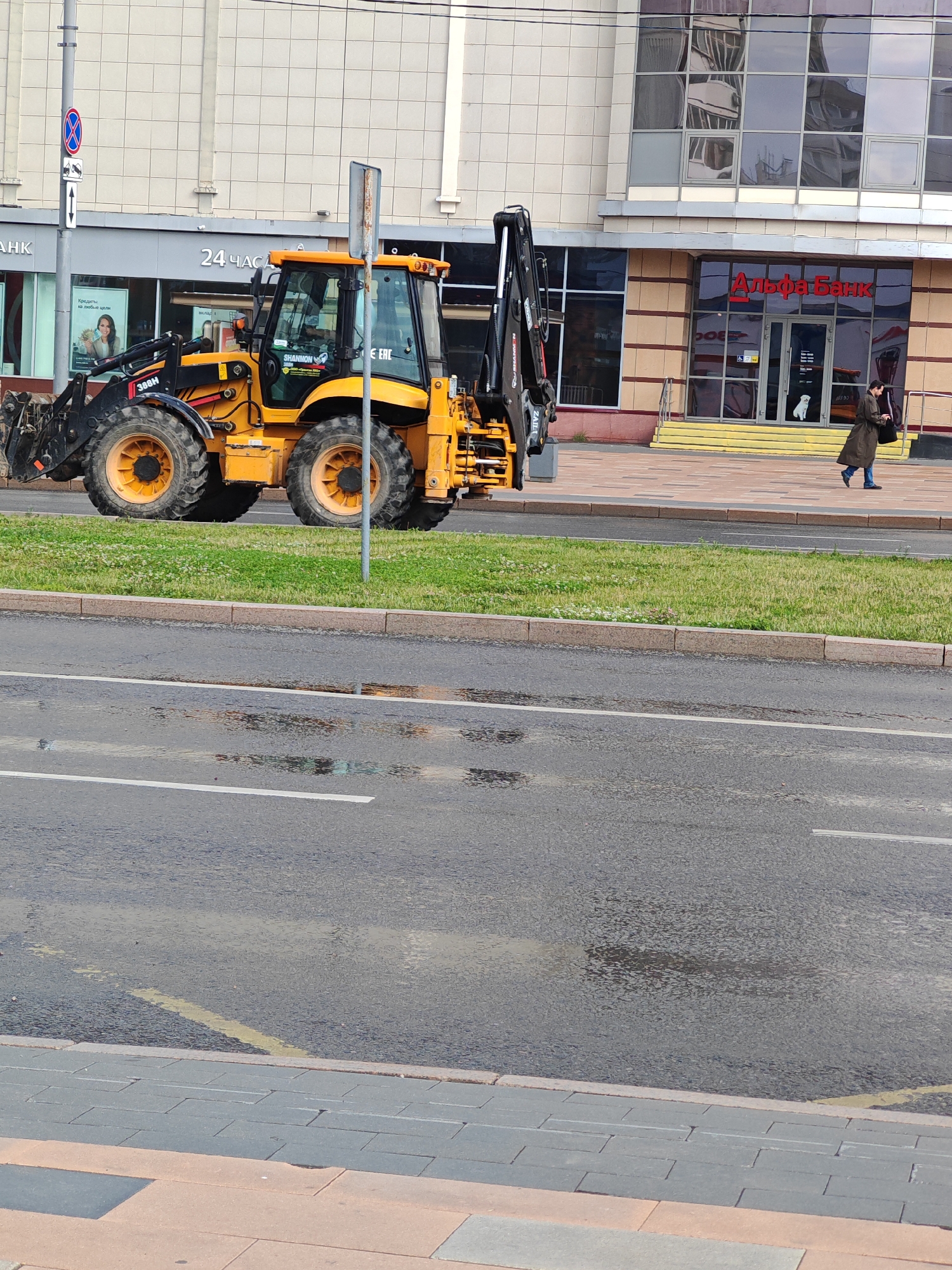 This media file shows a yellow backhoe loader parked on a grassy area next to a street. The backhoe is a large piece of construction equipment with a bucket at the front and an excavator arm at the rear. In the background, there is a modern building with large glass windows. A woman in a dark coat is walking on the sidewalk in front of the building. The building appears to be a bank, as indicated by the text "Альфа-Банк" (Alfa-Bank) visible on the facade. There are also signs with text such as "24 ЧАСА" (24 hours) and "Кредиты на любые цели" (Loans for any purpose). The street in the foreground is wet, suggesting it may have rained recently or is about to rain. The sky is overcast, contributing to the subdued lighting. The scene appears to be set in an urban environment during the daytime. There are no other people clearly visible in the image besides the woman walking. The overall impression is one of a typical urban street scene with construction equipment and a commercial building.
