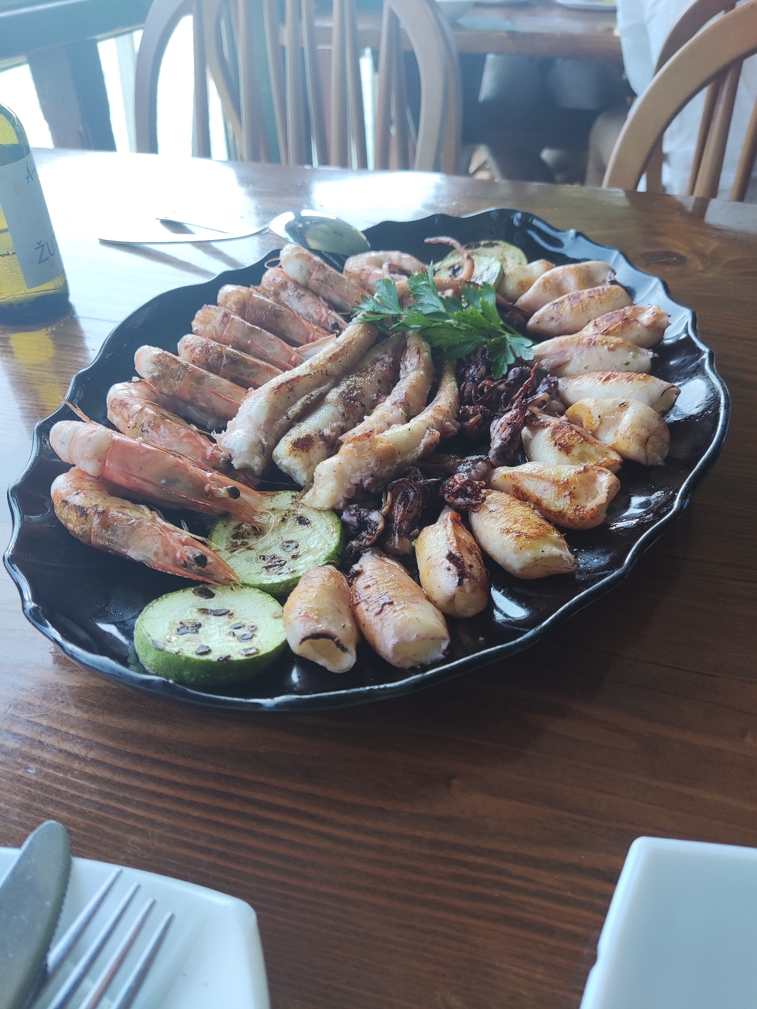 A black, wavy-edged platter filled with grilled seafood and zucchini slices sits on a dark wooden table. The platter contains several large, grilled shrimp with visible heads and tails, as well as numerous pieces of grilled squid, some whole and some cut. Grilled zucchini slices, round and green with char marks, are interspersed among the seafood. A sprig of fresh parsley garnishes the center of the platter. To the left of the platter, a partial view of a bottle of white wine with a label is visible. In the foreground, a white rectangular plate holds a fork and knife. The background shows the blurred shapes of wooden chairs and a window with bright light, suggesting an indoor dining setting. The scene is consistent with a meal at a restaurant in Gornji Štoj - Shtoji i Epërm, Montenegro.