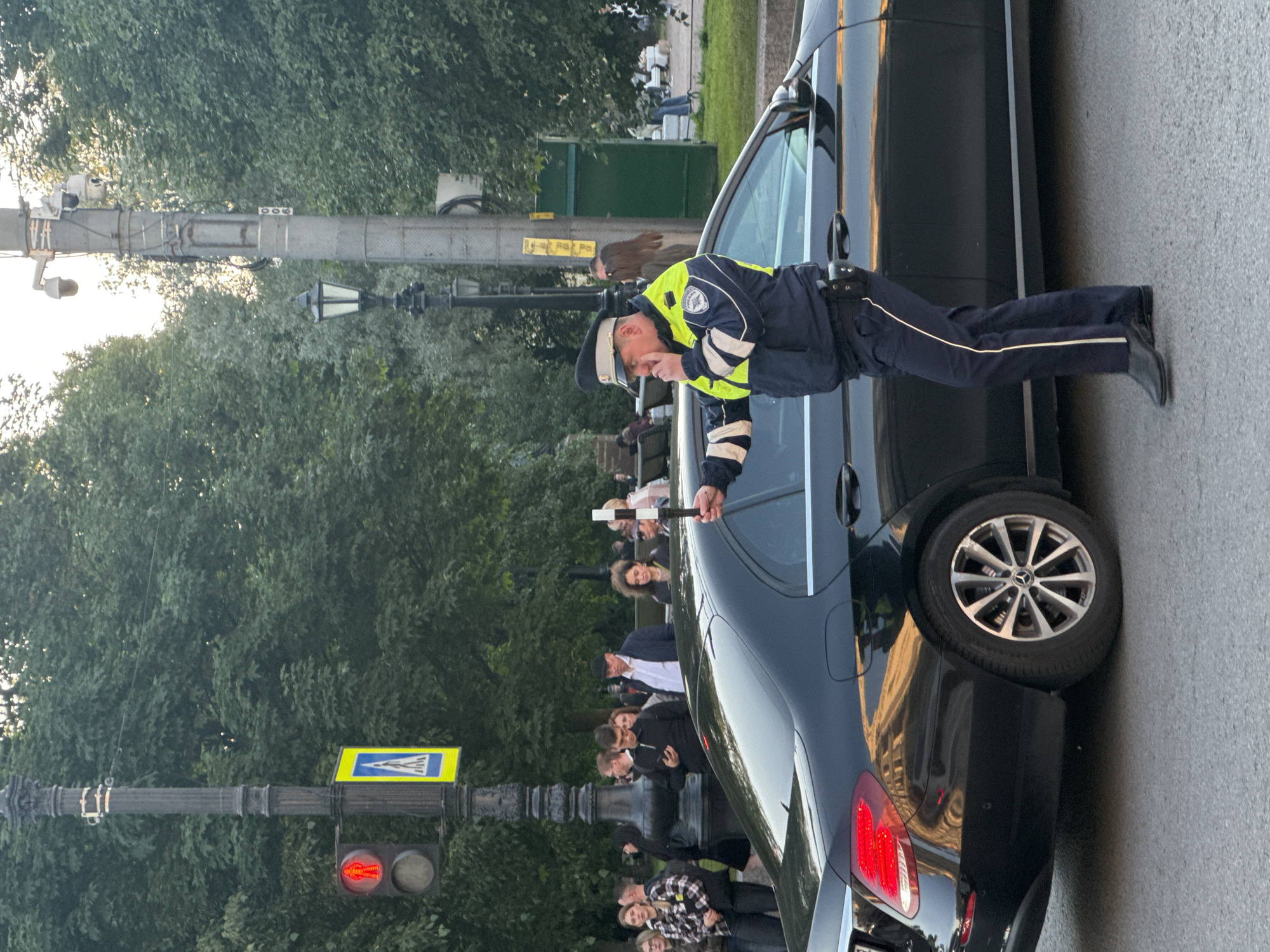 In Saint Petersburg, Russia, a police officer in uniform, wearing a dark blue jacket with high-visibility yellow and white accents and dark blue trousers, stands on a paved road next to a black Mercedes-Benz E-Class sedan. The officer is holding a white baton in their right hand and appears to be speaking into a communication device in their left. Behind the officer, a diverse group of pedestrians are visible on a sidewalk and grass area. One woman, wearing a plaid shirt, is in the foreground on the left, looking towards the viewer. Further back, several other individuals are standing and walking, with trees forming a dense green backdrop. A traffic light displaying a red signal is visible on a lamppost to the left, next to a pedestrian crossing sign. The overall lighting suggests it is late afternoon or early evening.
