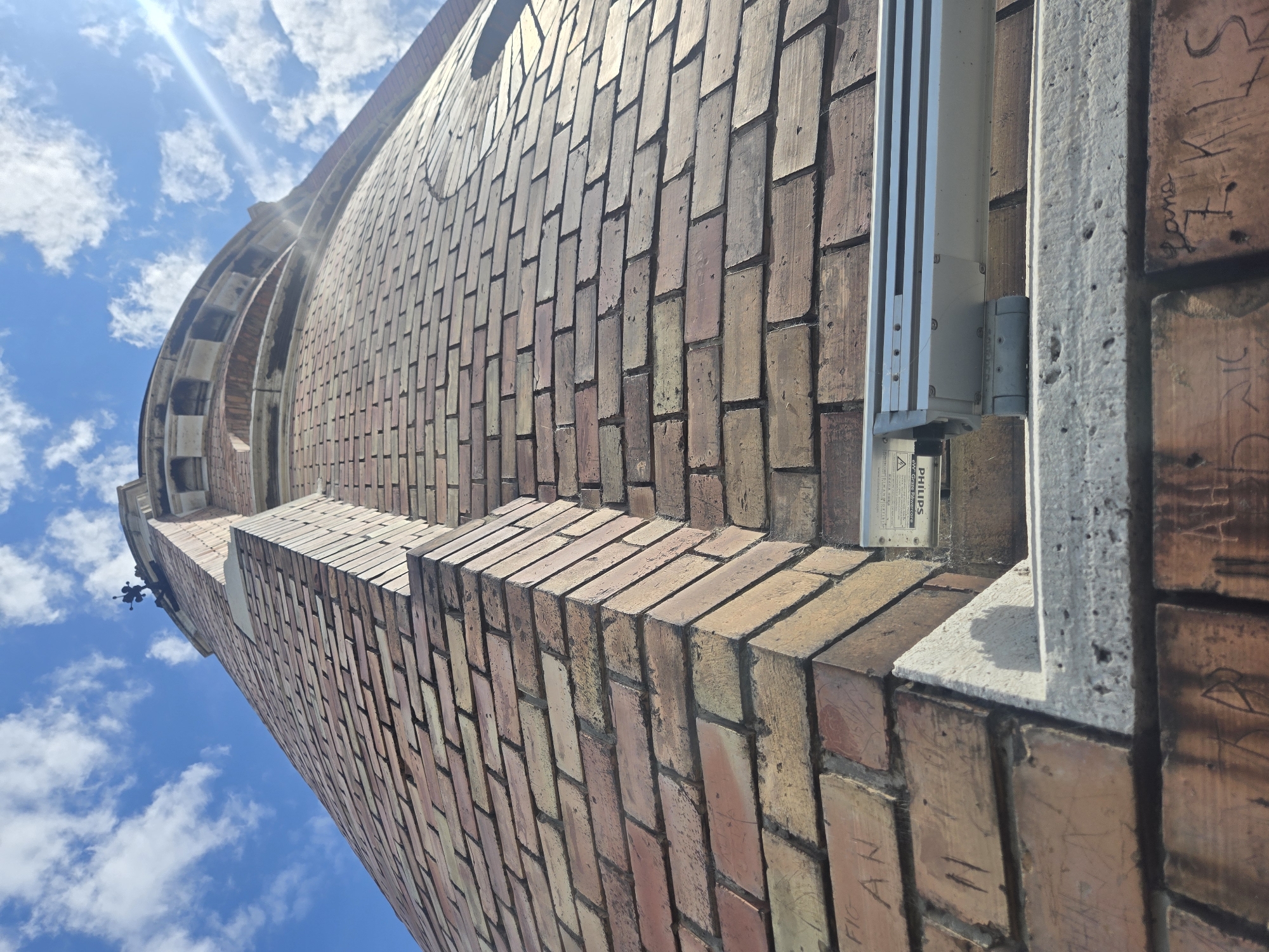 A low-angle shot depicts a brick structure under a blue sky with scattered clouds. The brickwork is primarily red and brown, with some graffiti present on the bricks closest to the right edge. A gray, metallic object, possibly a fixture or piece of equipment, is mounted on the wall. The structure appears to be cylindrical near the top left, curving upwards towards the sky. Given the architectural style and the graffiti, the location is most likely in Zemun Urban Municipality, Serbia.