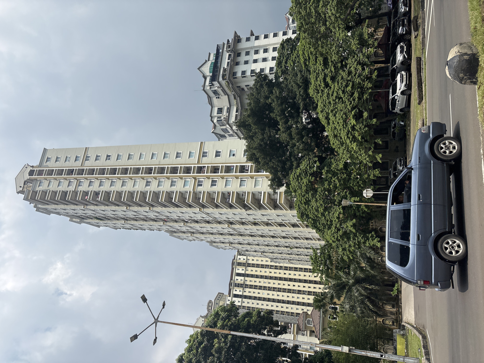 The image shows a street scene in Central Jakarta, Indonesia. A blue car is parked on the right side of the road. Along the street, several parked cars are visible under the shade of large trees. A tall, light-colored apartment building rises in the background, with a second apartment building also visible. Street lights are present. The sky is overcast.