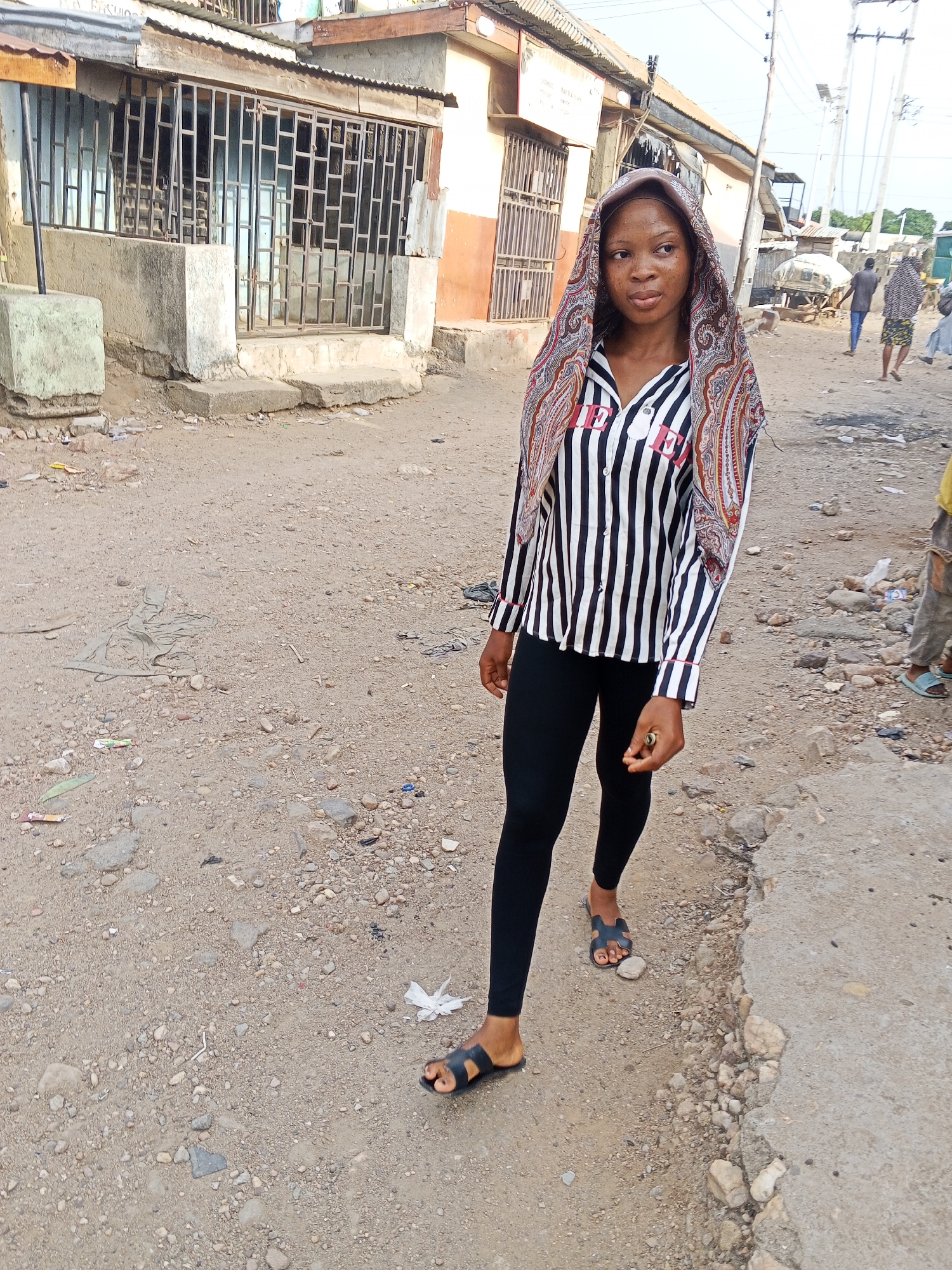 A young woman stands on a dirt and concrete road in Adankolo, Nigeria. She wears black leggings, black sandals, and a black and white striped shirt with a patterned headscarf. Behind her, buildings with metal bars on the windows line the street. Debris litters the ground. In the distance, people are walking. Power lines are visible in the sky.