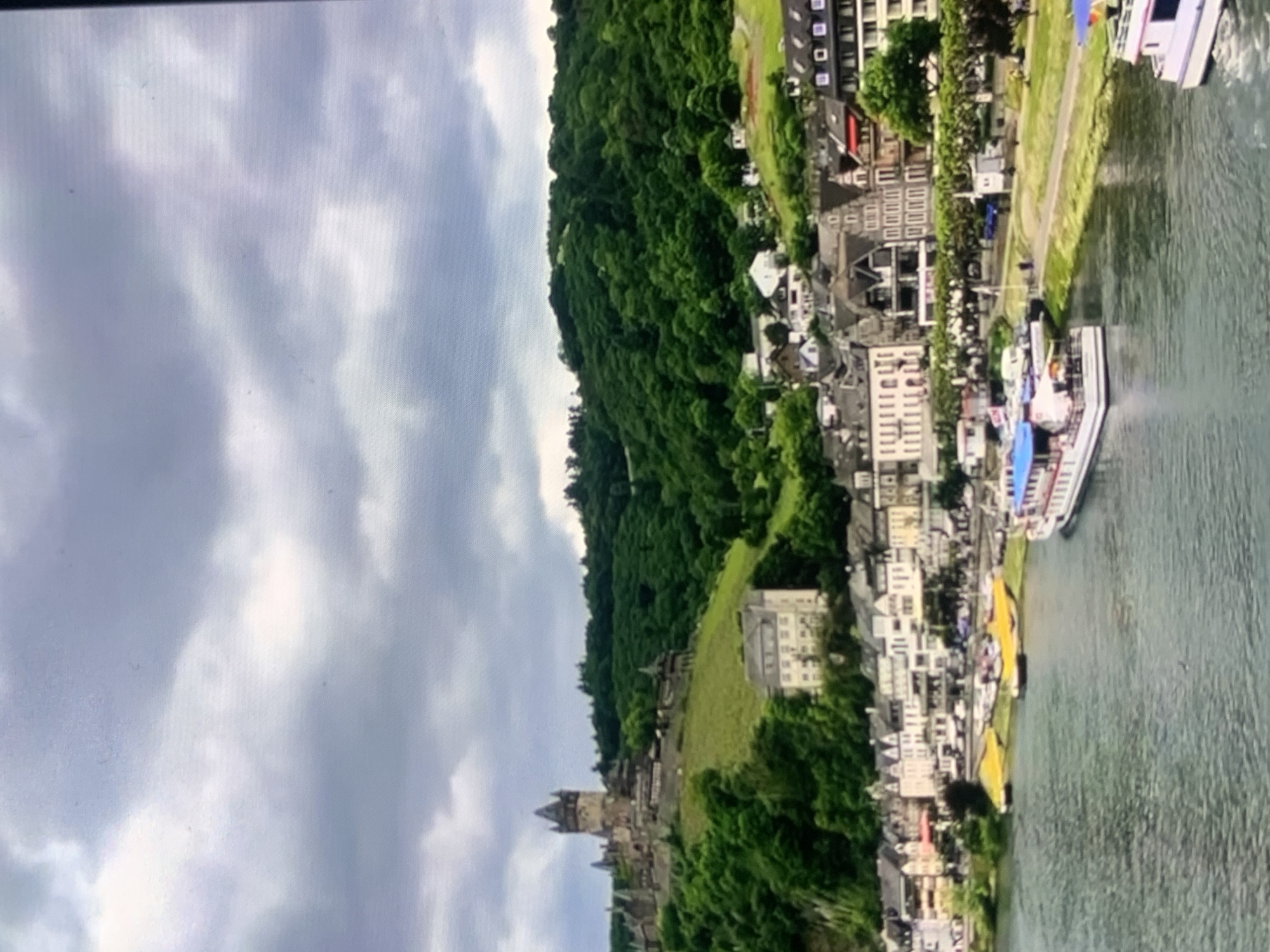 The image depicts a scenic European town situated on a riverbank, with a castle perched on a hill overlooking the scene. The town features traditional architecture with buildings in varying shades of white, grey, and red. A river flows in the foreground, with several boats docked or sailing on it. The background is dominated by a lush green hillside covered in trees, leading up to the prominent castle with its distinctive towers. The sky is overcast with grey clouds. There are no recognizable landmarks or elements that would specifically place this scene within the city of Stavropol, Russia.