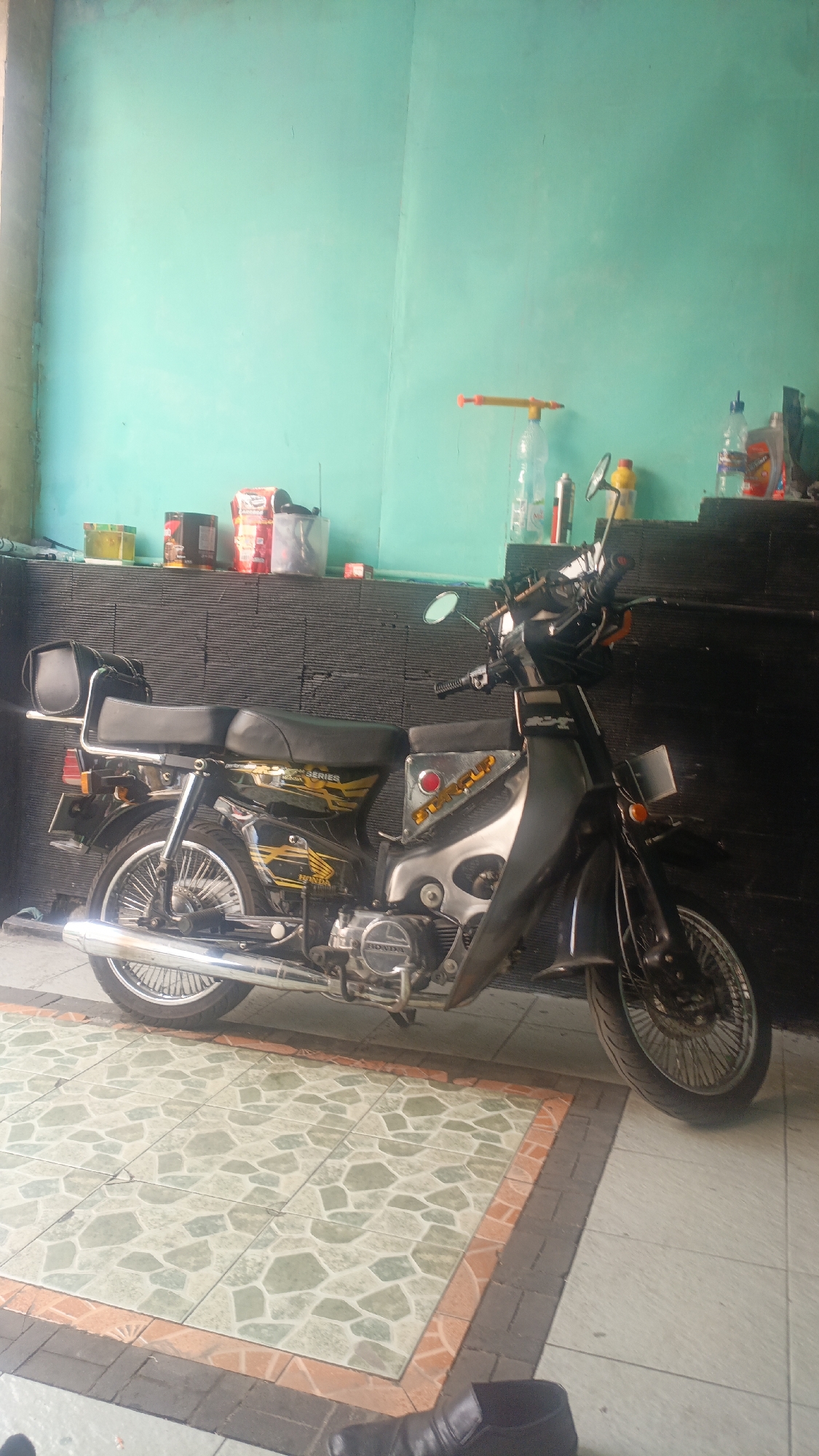 A motorcycle is parked in front of a teal-colored wall with a black textured facade behind it. The motorcycle is black with yellow and white decals, including the words "HONDA" and "STAR CUP SERIES". It has a black seat, a chrome exhaust pipe, and spoked wheels. On a shelf above the facade are several items including a water bottle with a red pump attachment, a yellow bottle, and various containers. The ground in front of the motorcycle is tiled with a green and white geometric pattern, bordered by orange and grey bricks. A black leather shoe is visible in the lower right corner of the image. The scene appears to be outdoors, possibly a porch or garage, in Salatiga, Indonesia.