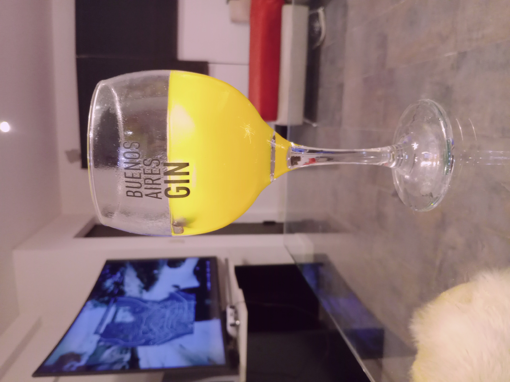 A yellow liquid is in a clear gin glass. The words "Buenos Aires Gin" are printed on the glass. The glass is on a glass table in a modern living room. A television is visible in the background.