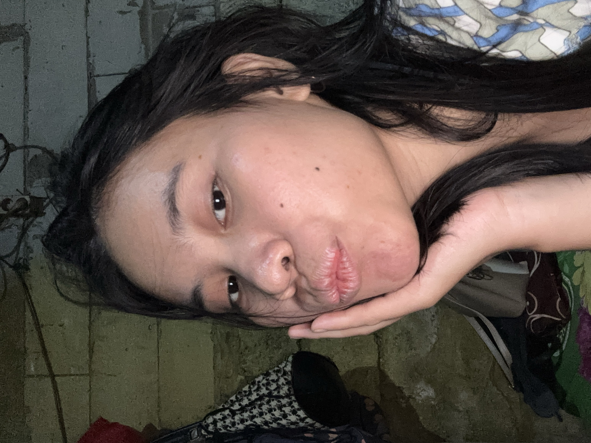 The media file is a close-up, vertically oriented photo of a young person's face, tilted to the side. The subject has long, dark hair framing their face and is resting their head on their hand. Their lips are puckered, and their eyes are open, looking towards the camera with a neutral expression. There are a few small moles visible on their cheek.
The background is dimly lit and appears to be an indoor setting with textured, possibly concrete, walls and some scattered objects, including patterned fabric, bags, and clothing. The lighting suggests it is likely nighttime or a very dark indoor environment. There are no visible signs or text in the image. The overall mood of the image is intimate and perhaps slightly melancholic due to the subject's pose and the subdued lighting. The location context is East Jakarta, Indonesia.
