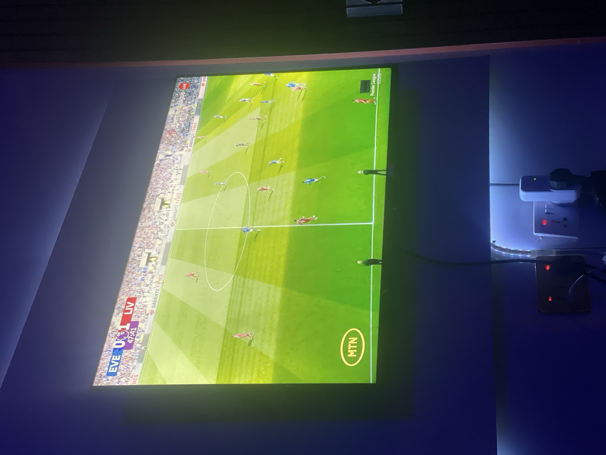 The image shows a television screen displaying a live broadcast of a Premier League football match between Everton (EVE) and Liverpool (LIV). The score is 0-1 to Liverpool, with the time at 47:41. The MTN logo is visible, indicating the broadcast is likely from Africa. The screen is mounted on a dark blue wall, next to an electrical outlet with multiple devices plugged in. The broadcast includes advertising for Davanti Tyres and shows a general stadium view with players on the field. There is no indication of the city of Lokoja, Nigeria, in the image.