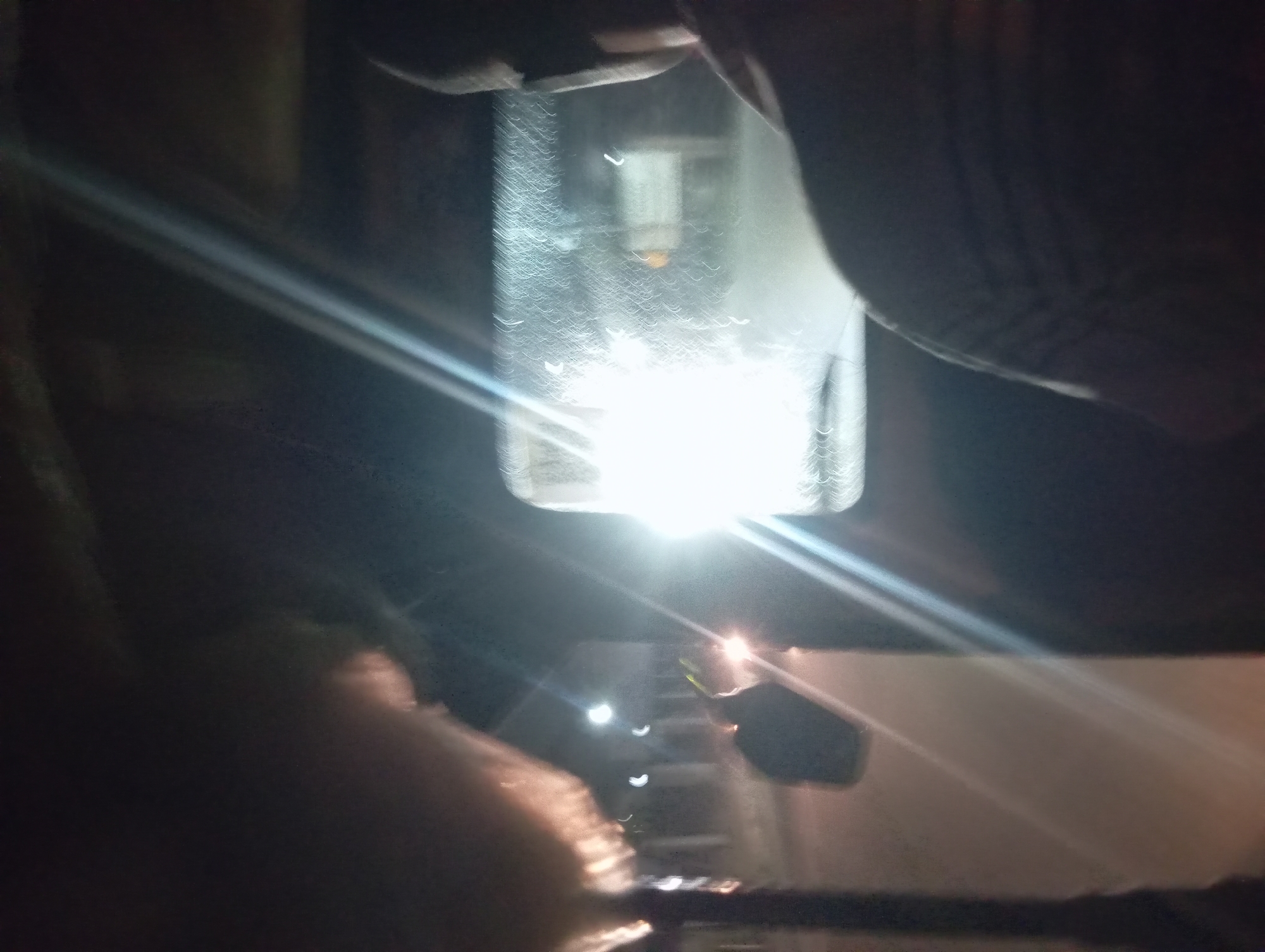 The image depicts a nighttime scene from inside a moving vehicle, likely a rickshaw given the context of Onitsha, Nigeria. The perspective is from a passenger looking forward through the windshield and towards the right side of the vehicle. The interior of the vehicle is dark, with the exception of the bright headlights of another vehicle coming towards them, causing lens flare and obscuring details on the windshield.
A person's head and shoulder are visible in the foreground on the right, their face obscured by darkness and the angle. They are wearing a striped shirt. In the mid-ground, through the windshield, the bright headlights of an oncoming vehicle are the dominant feature. Streetlights and the blurry outlines of buildings and possibly other vehicles are faintly visible in the background. On the left side of the image, a fluffy, light-colored object, possibly a piece of padding or fabric, is in the upper left corner, partially obstructing the view. To the left and slightly behind the front of the vehicle, a dark-colored object that appears to be another vehicle is visible. The overall impression is of a journey at night on a street in Onitsha, Nigeria, with limited visibility due to darkness and the bright headlights of approaching traffic.