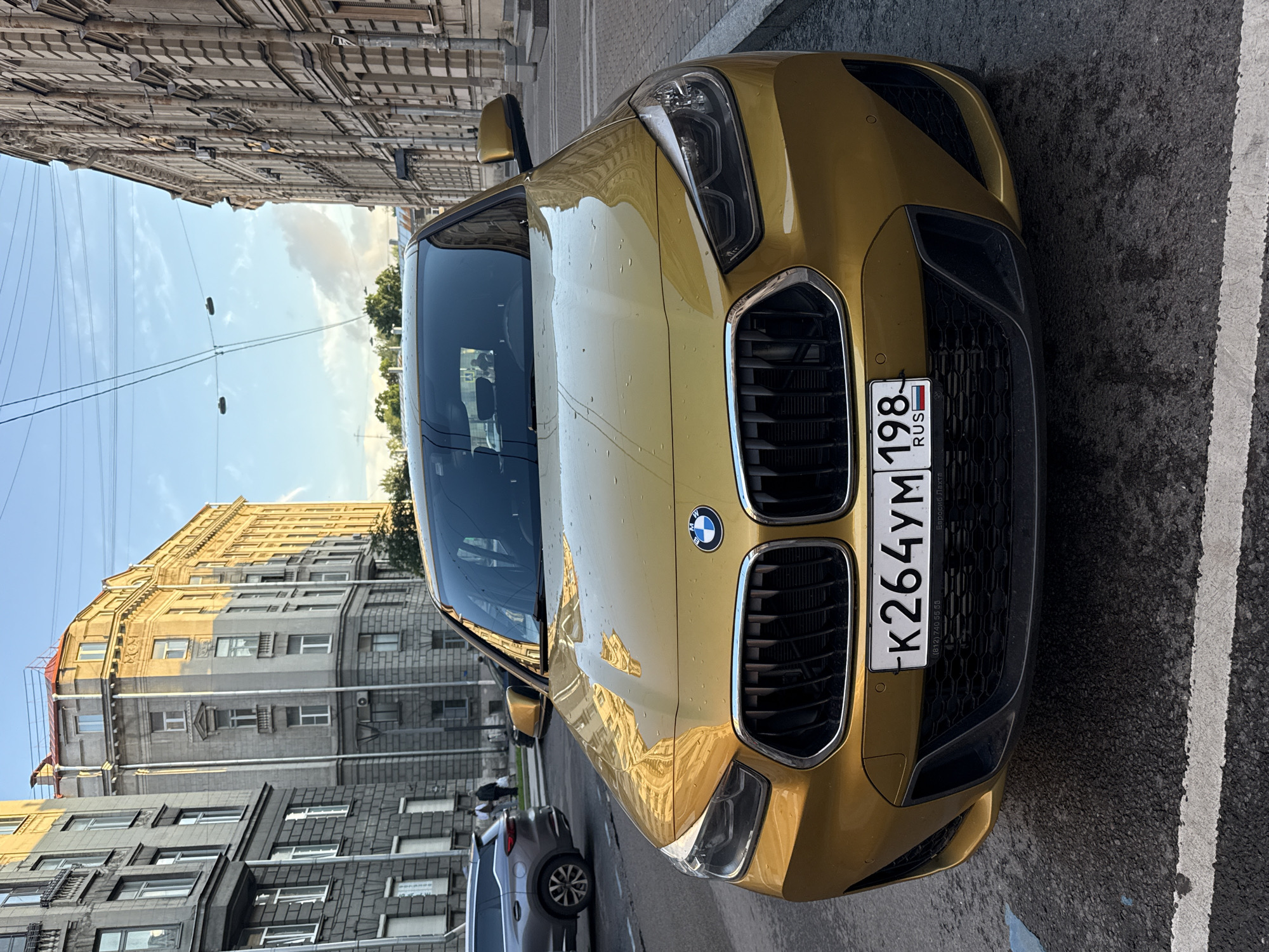 A golden BMW X2 is parked on a city street in Saint Petersburg, Russia. The car is facing the camera, with its headlights on and its license plate visible. The license plate reads "K264YM 198 RUS". The street is paved and there are buildings on both sides of the street. The sky is blue with some clouds. The buildings appear to be old, with ornate architecture, characteristic of Saint Petersburg. One of the buildings on the left has several windows with closed blinds. On the right, another building shows air conditioning units mounted on its facade. In the background, a grey SUV is parked on the left side of the street. The overall scene depicts a typical urban environment in Saint Petersburg, Russia, with a focus on the BMW X2.