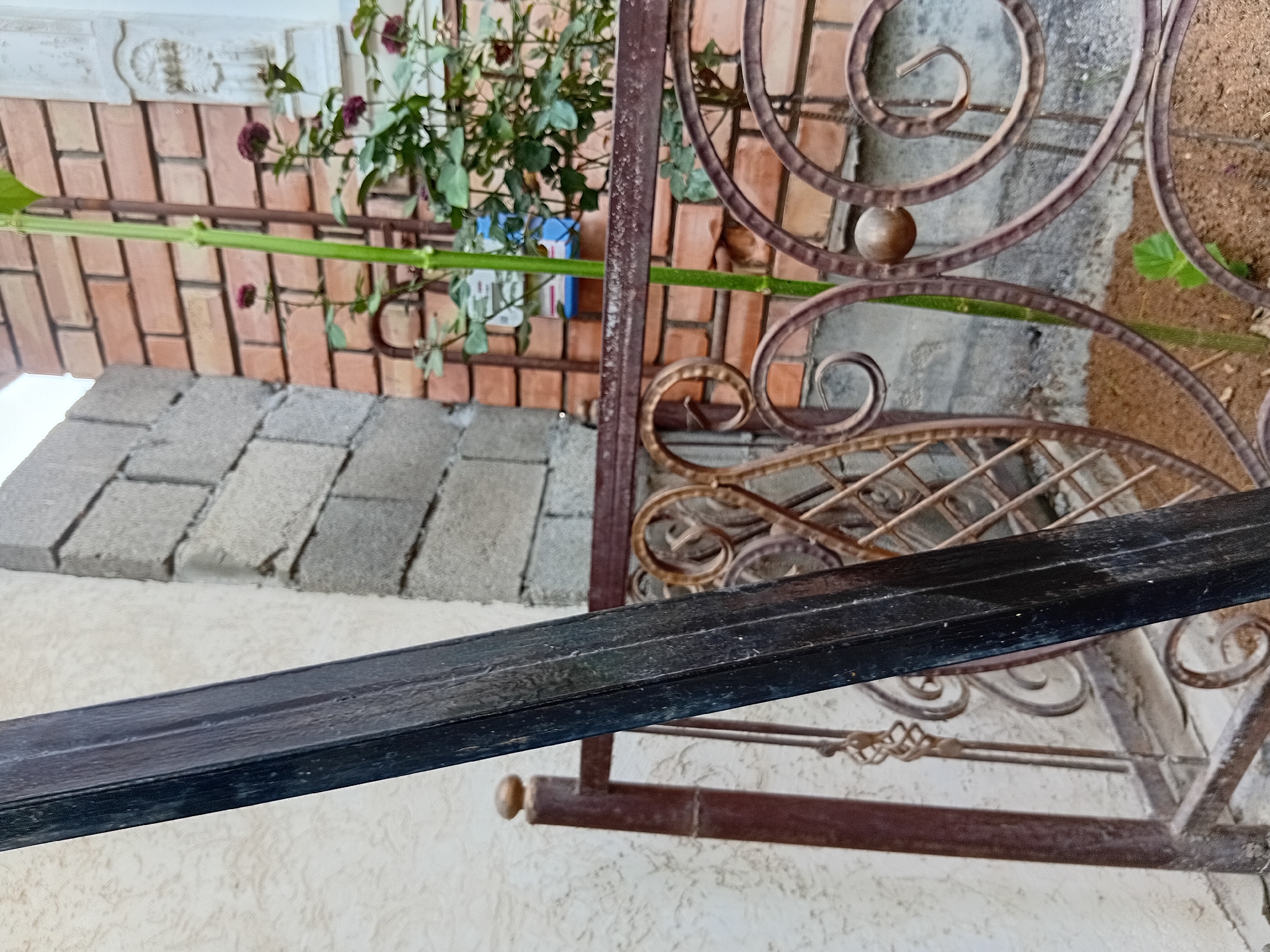 The image is a close-up shot of a section of a yard in Kadamjay, Kyrgyzstan. In the foreground, a dark, weathered metal beam is angled diagonally from the left side of the frame towards the bottom right. Behind the beam, there is decorative wrought iron fencing with circular and scrollwork patterns. A single green stem from a plant with small red flowers grows up through the fencing. The background consists of a textured, light-colored wall on the left, a rough concrete block wall in the middle, and a brick wall on the right. A light-colored decorative element is visible on the top right of the brick wall. The ground beneath the fencing appears to be covered in loose soil or sand.