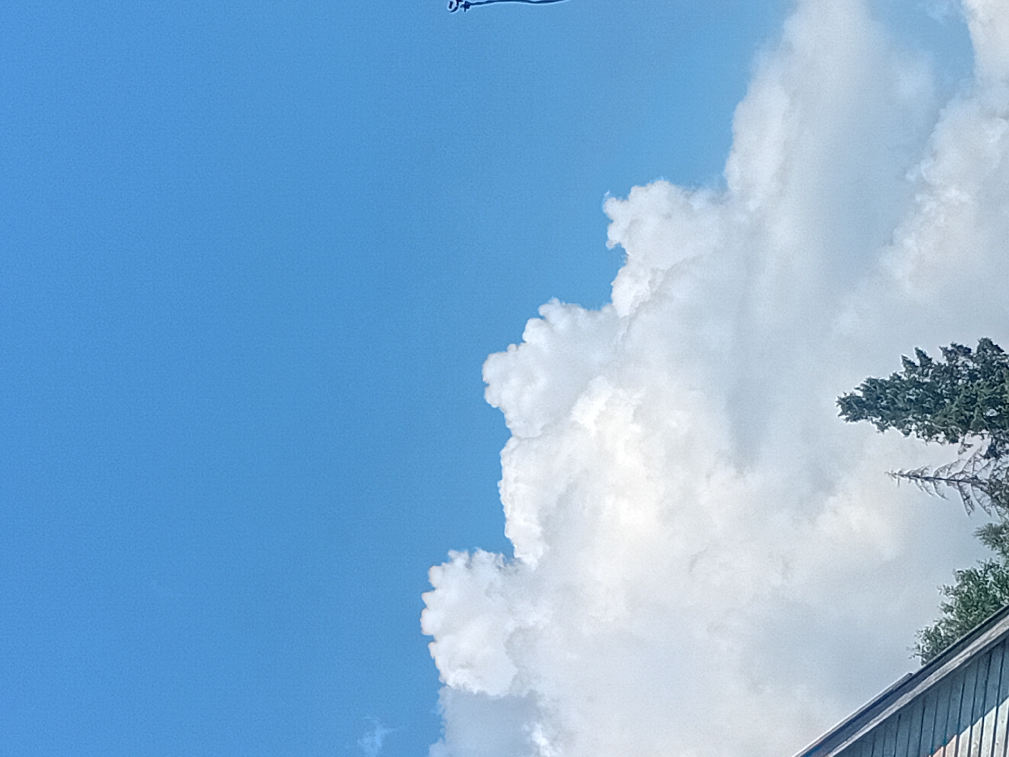 The image depicts a clear blue sky with scattered white cumulus clouds. In the lower left portion of the frame, the roof and side of a building are visible, constructed with a dark-colored material, possibly wood shingles. To the right of the building, several tall evergreen trees are present, with one notably sparser and lighter in color than the others. On the far right side of the image, a portion of a dark pole or mast is visible extending upwards into the sky, with a small blue object attached near its top. There are no people or animals present in the scene. The overall impression is a daytime, outdoor scene, likely in a rural or suburban area given the presence of trees and a building. The location is specified as Verkhniachka, Ukraine.