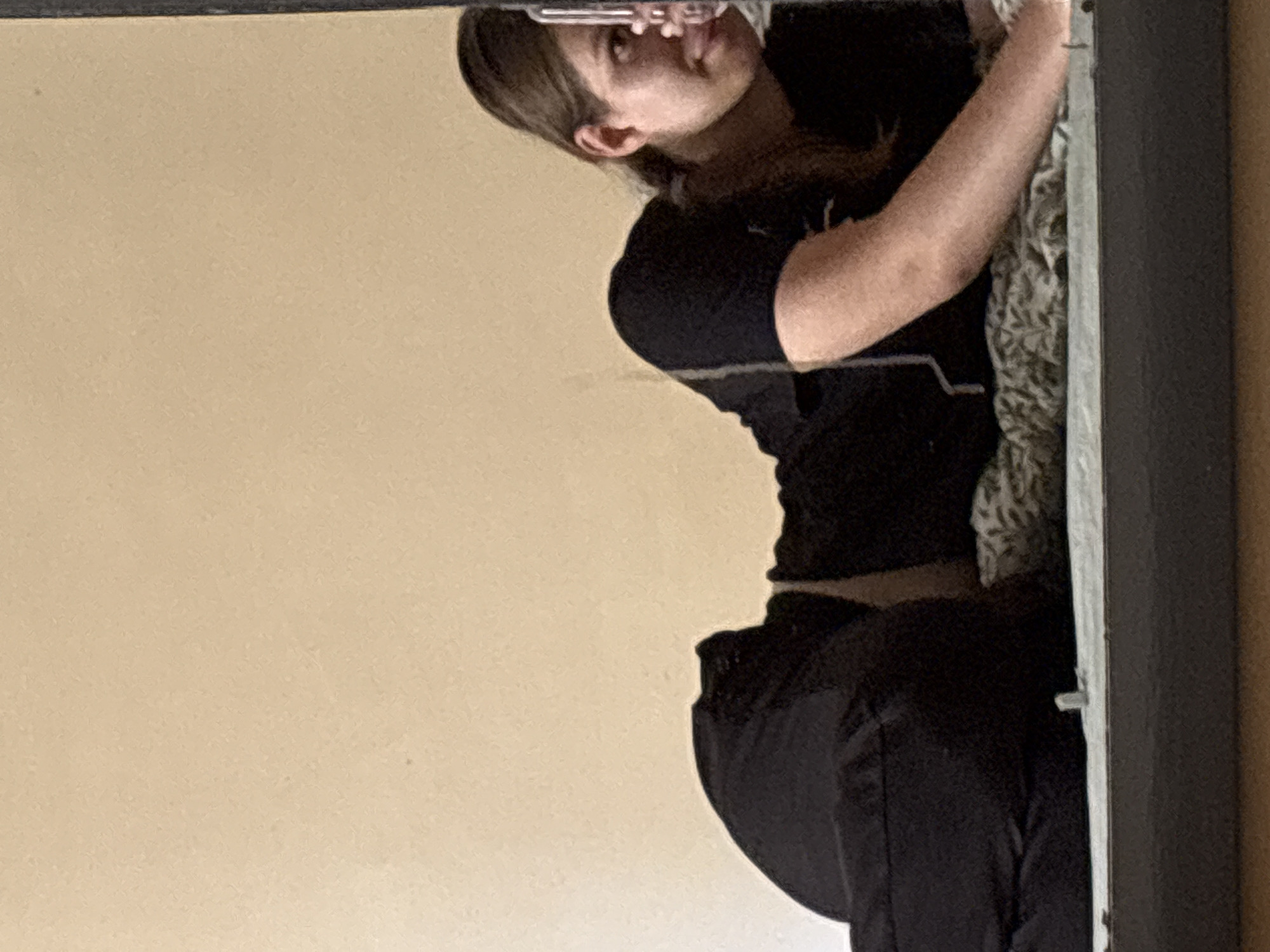 A reflection in a mirror shows a woman in a black top and pants holding a mobile phone. A wall appears in the reflection, as does the edge of what appears to be a bed or sofa. The mirror has a dark frame. The location is not discernable from the image.