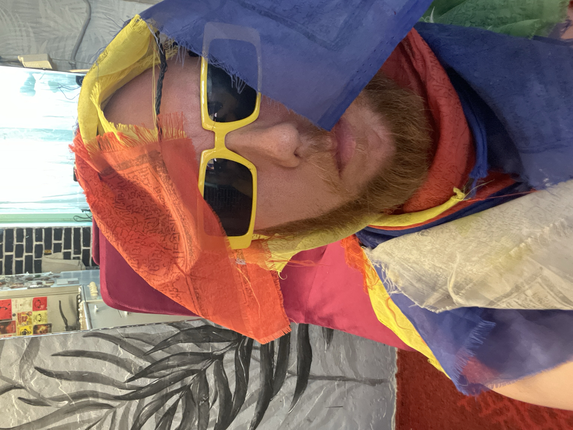 In Saint Petersburg, Russia, a person with a ginger beard and wearing bright yellow rectangular sunglasses is captured in a close-up, head-and-shoulders shot. They are draped in colorful prayer flags, predominantly red, yellow, blue, and white, which obscure much of their face and head. The flags have a frayed, tasseled edge and appear to be made of a lightweight fabric. The individual is seated in a bright pink chair. In the background, a grey and black leaf-patterned tapestry hangs on the wall to the left. To the right, a brick wall with a green curtain is visible. The overall impression is of a vibrant and eclectic indoor setting.