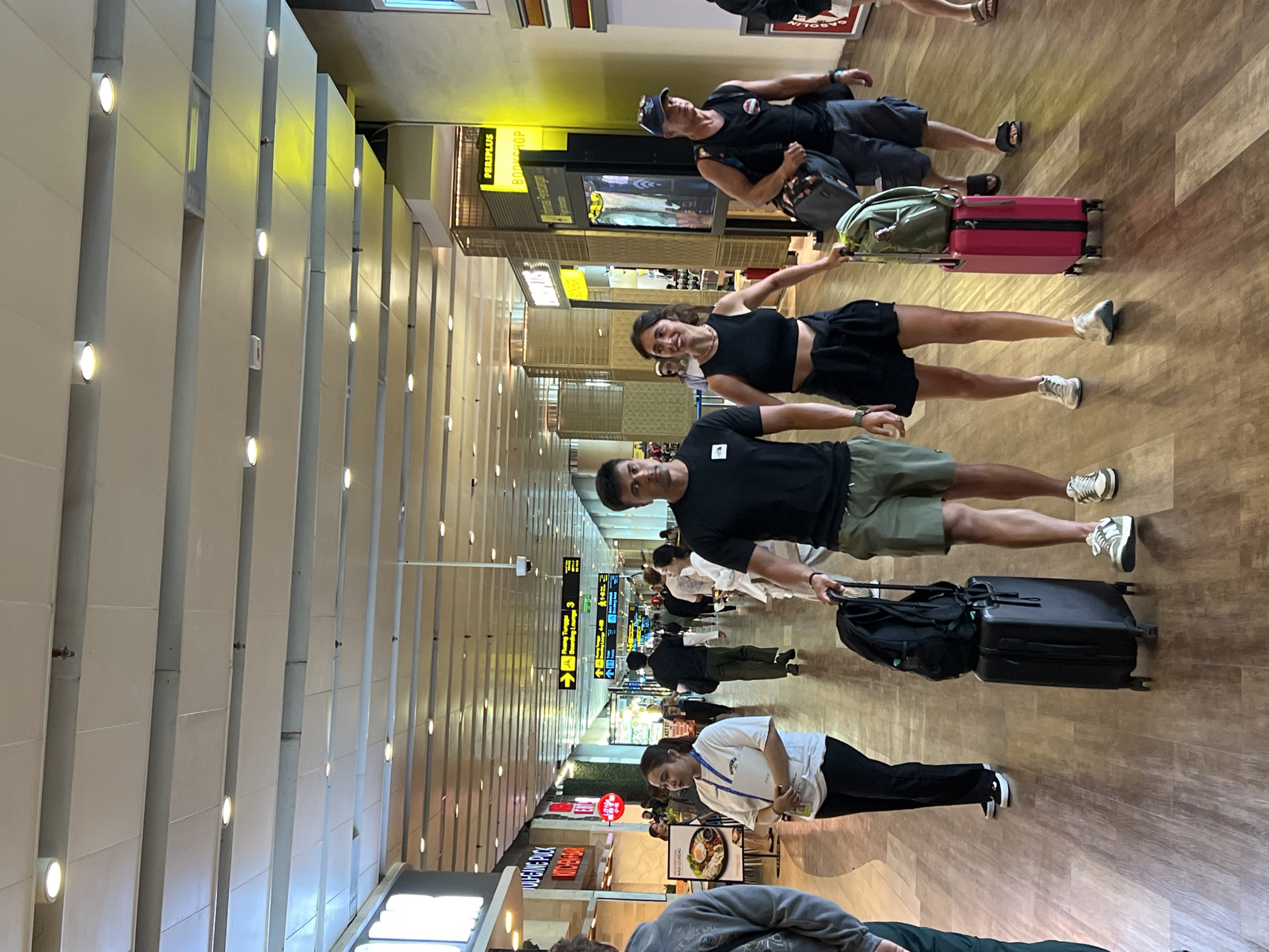 The image depicts a busy airport terminal, likely at Ngurah Rai International Airport in Kuta, Indonesia. Several individuals are walking down a wide, well-lit corridor with shops and signage visible on either side. The floor is made of polished wood. In the foreground, a couple is walking together, the man on the left is wearing a black t-shirt and olive green shorts, and he is pulling a black suitcase with a black backpack on top. The woman next to him is wearing a black crop top and black shorts, and she is pulling a pink suitcase with a green backpack on top. Other travelers are seen in the background, some with luggage, indicating movement and transit. The atmosphere appears to be one of typical airport activity.