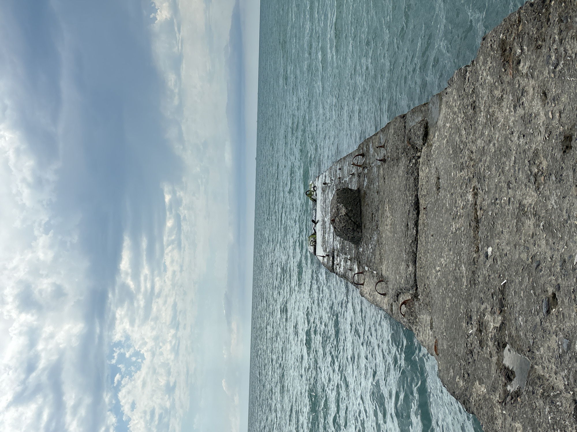 This media file depicts a wide, choppy expanse of pale blue-green water under a cloudy sky. A concrete structure, likely a pier or breakwater, juts out from the right side of the frame, its rough texture contrasting with the smooth water. The concrete is weathered and shows signs of reinforcement, with several rusty metal loops protruding from its surface. A large, rounded stone is embedded in the concrete near the end of the structure. A few small birds, possibly seagulls, are perched on the concrete. The scene is set in Sochi, Russia, and the lighting suggests it is daytime, with the clouds indicating overcast or partly cloudy weather. The overall mood is one of natural elements interacting, with the man-made structure a sturdy presence against the dynamic water and sky. No text is visible in the image.