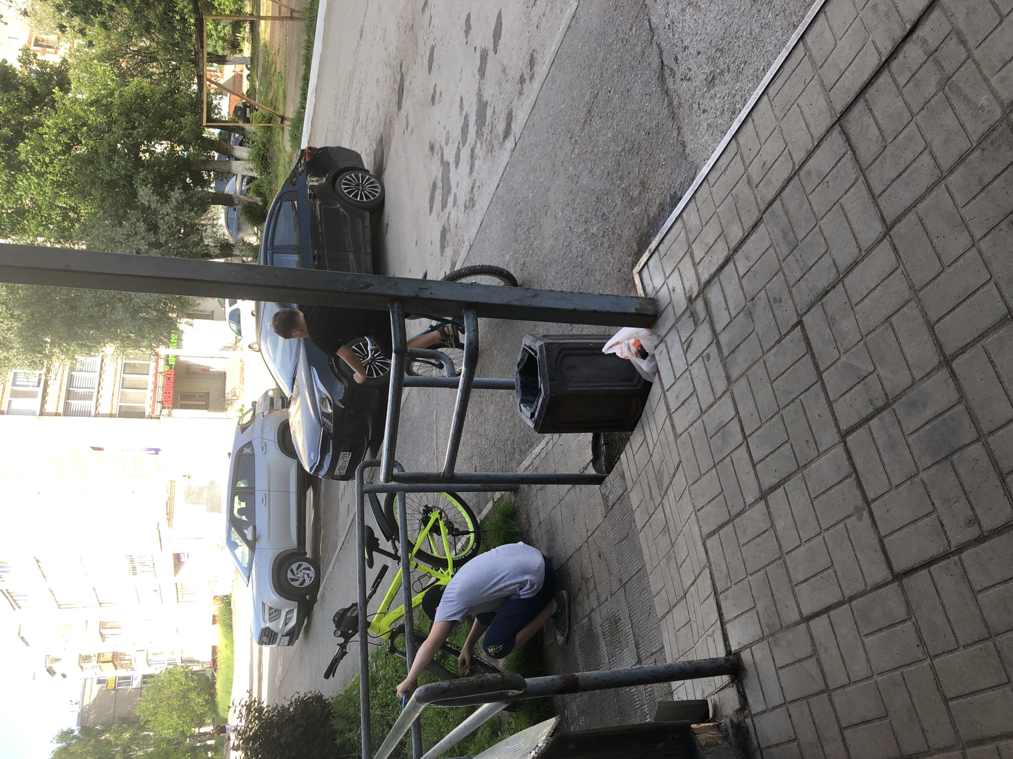 A young male is crouched on a paved walkway, attempting to repair a bright yellow bicycle. He is wearing a grey t-shirt and dark shorts, with grey sneakers. The bicycle is leaning against a metal railing, with its front wheel near the male. In the background, a black sedan is parked on an asphalt street, with a silver SUV parked further down the street. The scene appears to be in a residential area, with apartment buildings visible in the distance. A black trash bin is located to the right of the walkway. This location is in Solikamsk, Russia.