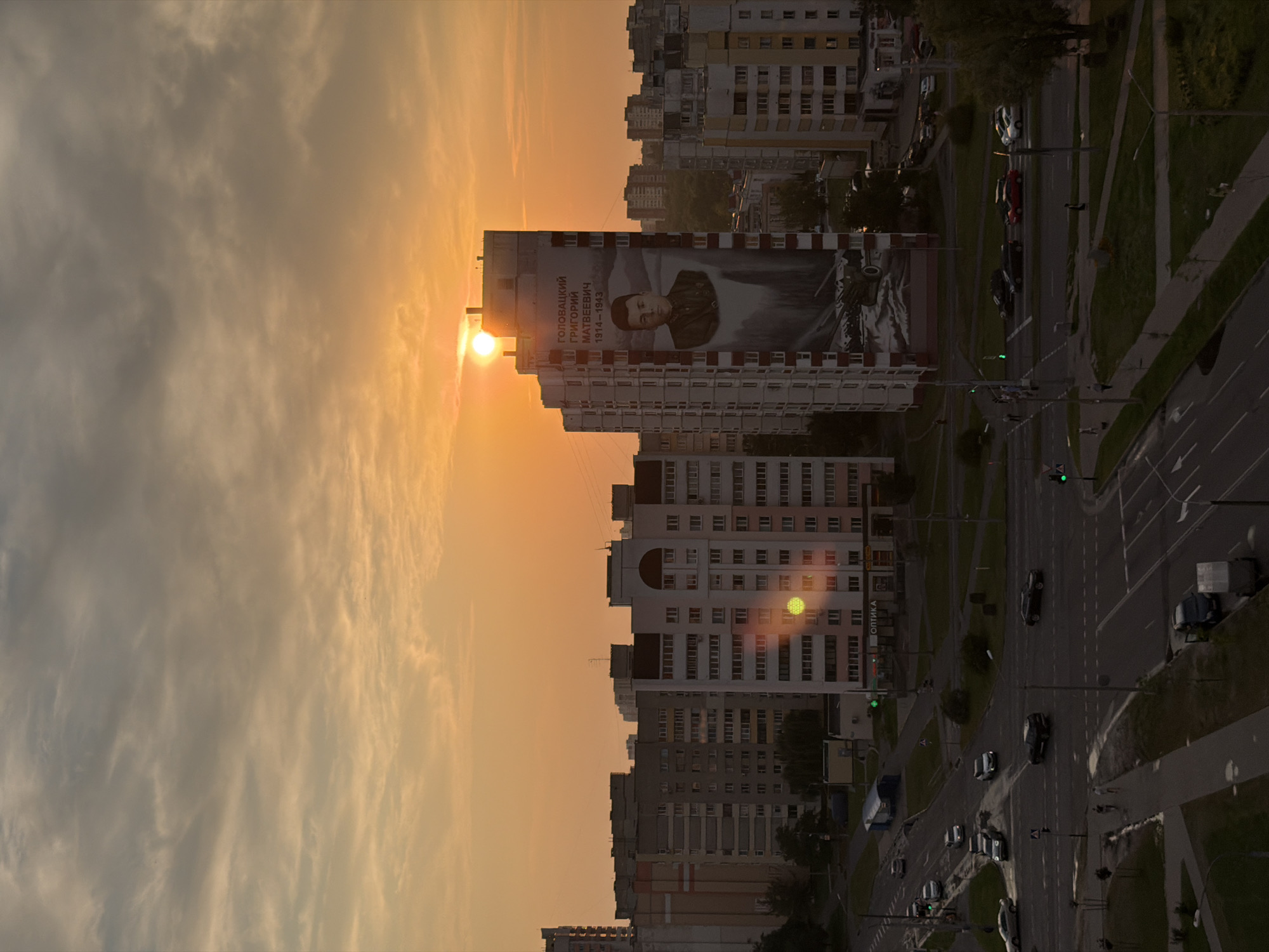 The image depicts a view of a residential area in the city of Homyel, Belarus, during sunset. The sun, a bright orb, is partially obscured by clouds and hangs low in the sky, casting a warm, orange glow over the scene. In the foreground, a multi-lane road with several cars is visible, some appearing to be in motion. Pedestrians can be seen walking on sidewalks. The middle ground features a series of apartment buildings, typical of Soviet-era architecture, characterized by their uniform design and multi-story structures. One prominent building displays a large mural of a man, identified by text above as "ГОЛОВАЦКИЙ ГРИГОРИЙ МАТВЕЕВИЧ 1914-1943". This mural is a significant focal point. In the background, more apartment buildings and trees can be seen, creating a dense urban landscape. The overall atmosphere is one of twilight, with the fading sunlight illuminating the city and casting long shadows.