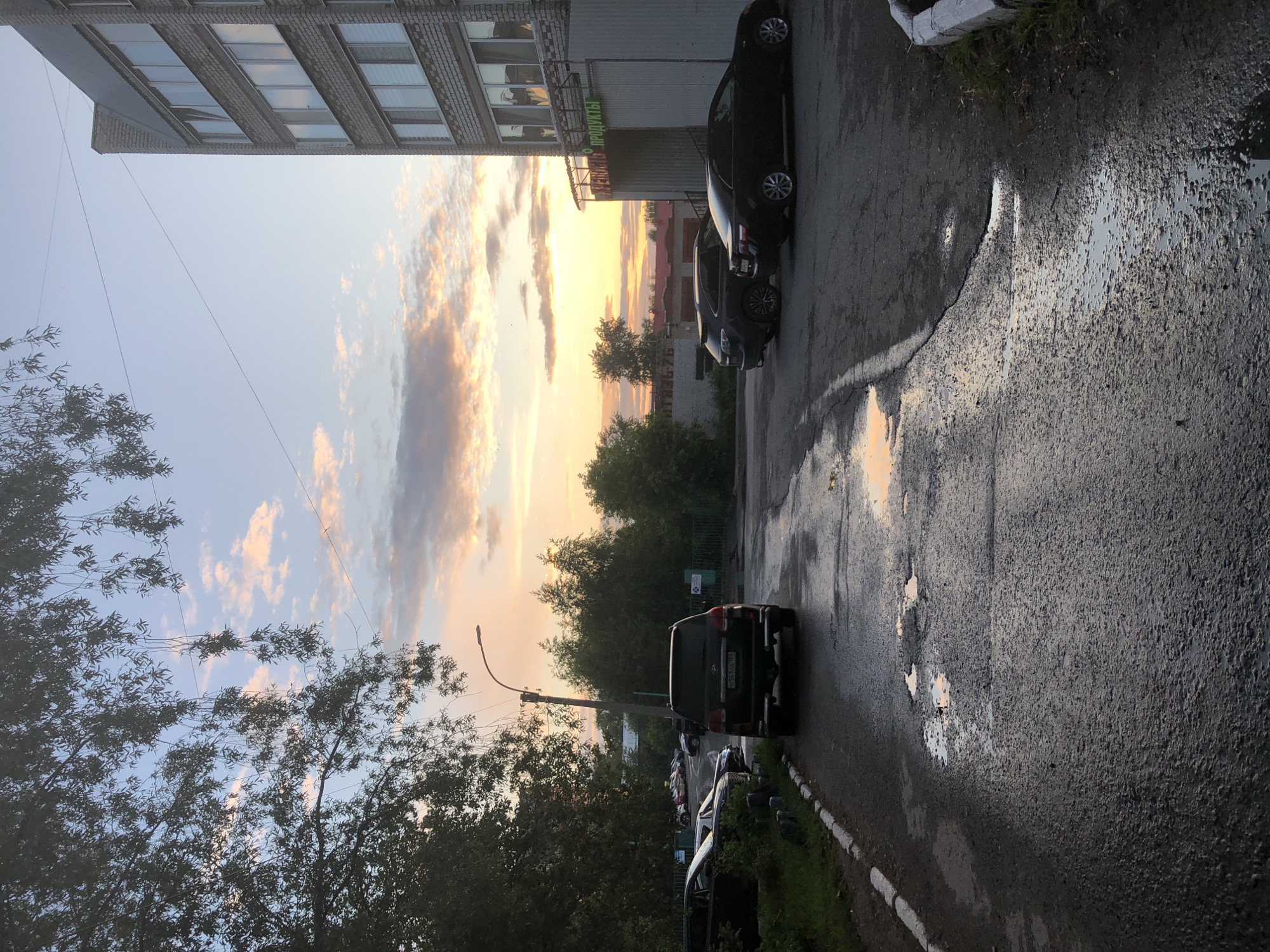The image depicts a street scene in Solikamsk, Russia, at dusk. A wet asphalt road, reflecting the colors of the sunset, dominates the foreground. Several cars are parked on the side of the road and in a parking lot. To the right, a multi-story brick building with many windows is visible, with a sign that appears to read "ПРОДУКТЫ" (Products). Trees and a street lamp line the left side of the road. The sky is a mix of blue and soft orange and pink clouds, indicating the sun is setting. The overall impression is of a quiet urban evening.