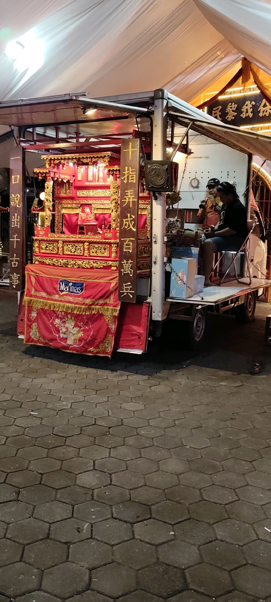 The image depicts an outdoor scene at night, likely in Pulo, Indonesia, with a street stall setup. The central focus is an ornate, red and gold altar or shrine mounted on a flatbed trailer. The structure is elaborately decorated with intricate carvings and golden embellishments, draped with red fabric and fringe. A banner with the "Marimas" logo is prominently displayed on the front.
To the right of the altar, two individuals are seated on the trailer. One person, wearing a dark shirt and shorts, is holding a phone. The other person, in an orange shirt and a cap, is playing a wind instrument. Behind them, there are boxes, speakers, and other equipment associated with a street performance or vendor setup.
The altar is flanked by tall wooden pillars with Chinese characters inscribed on them. The ground is paved with hexagonal grey interlocking stones. The scene is illuminated by artificial lights, casting shadows and highlighting the textures of the objects and the pavement. A white canopy extends over the setup, suggesting it's a temporary structure for an event or business. The overall impression is that of a lively street scene with a cultural or religious element.