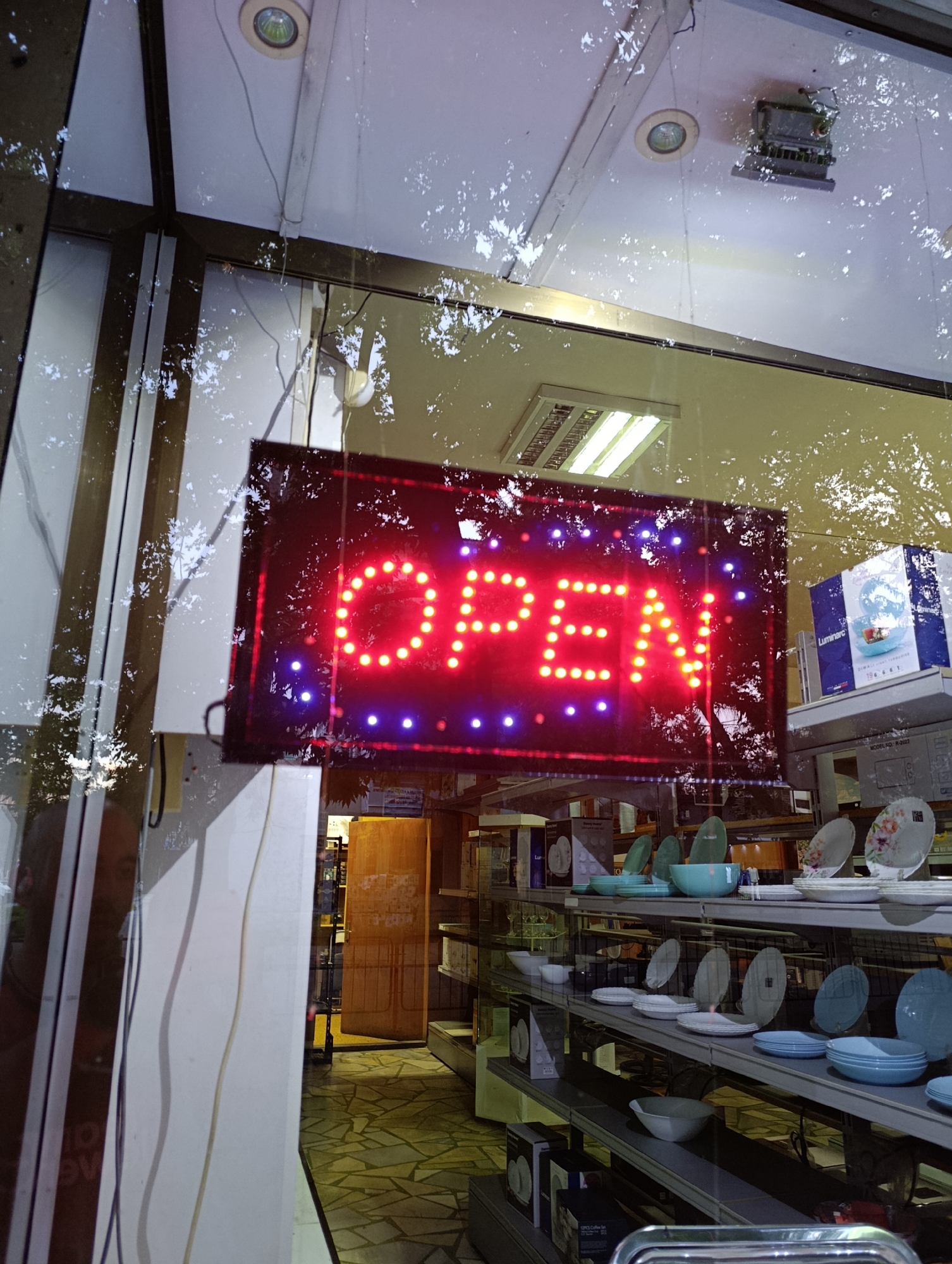 The image shows a close-up, slightly distorted view through a storefront window. A bright red neon sign spelling "OPEN" in all caps is the central focus, illuminated by yellow lights for the letters and purple lights along the bottom edge. The sign is framed by a dark border and appears to be hanging from wires.
Reflections on the window are prominent, showing trees, sky, and parts of the ceiling with lighting fixtures and electrical conduits, suggesting an outdoor environment. Inside the store, shelves are visible, stocked with various items including plates, bowls, and boxed goods, indicating a retail establishment, possibly a homeware or kitchen supply store. A wooden door stands ajar in the background, revealing more of the store's interior. The floor inside appears to be tiled. The overall impression is of a store that is open for business, with the neon sign clearly indicating its operational status.