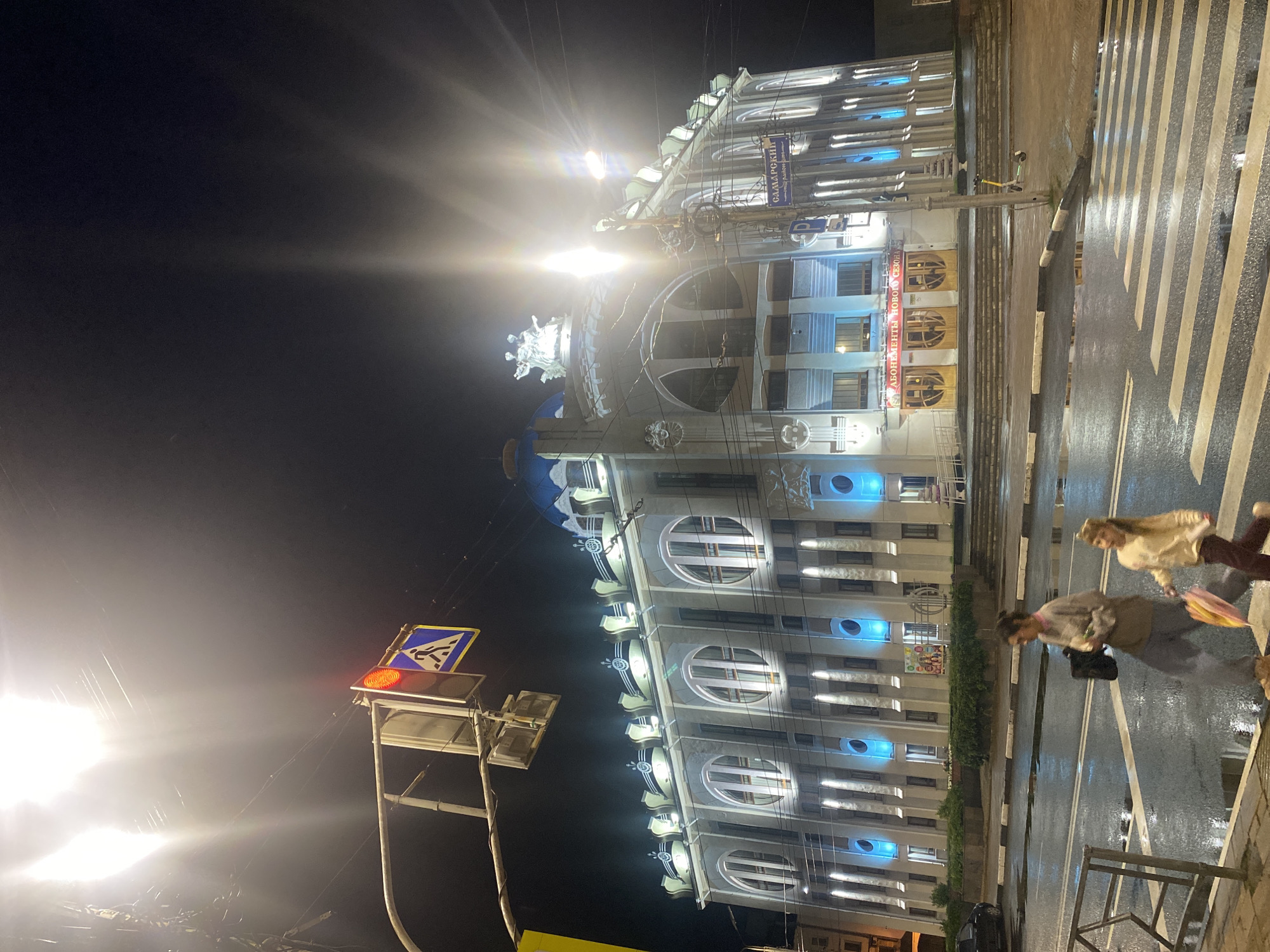A nighttime, low-angle shot captures the facade of a grand, illuminated building in Unknown, Unknown. The building, possibly a theater or cultural center, features a blue dome and an ornate facade with arched windows and decorative elements, all highlighted by white and blue lights. A streetlamp with intense glare dominates the upper left of the frame. A pedestrian crossing with zebra stripes is visible in the foreground, with wet asphalt indicating recent rain. Two females are crossing the street from left to right: an adult with dark hair wearing a grey sweatshirt and grey pants, carrying a black bag, and a younger female with blonde hair wearing a light-colored top and dark pants, holding a colorful umbrella. Traffic lights with a red signal are visible above the crossing. A street sign with Cyrillic lettering, possibly indicating a street name or district, is mounted on a pole near the building's entrance. An electric scooter is parked on the sidewalk to the right of the building.