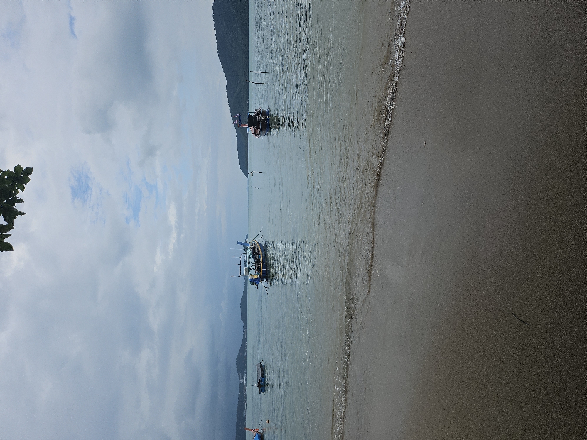 The image depicts a view of the sea and beach in Rawai, Thailand. In the foreground, wet sand shows a faint line of foam from an incoming wave. The water is calm and a light greyish-blue, with small waves lapping the shore. Several longtail boats are visible on the water. Two boats are anchored closer to the shore, one is a bright blue and yellow, and the other is smaller and darker with a tarp covering it. Further out, a red and blue longtail boat is anchored with two poles standing in the water nearby. In the distance, a hilly coastline with green trees is visible under a cloudy sky. The leaves of a plant hang from the top right corner of the frame.