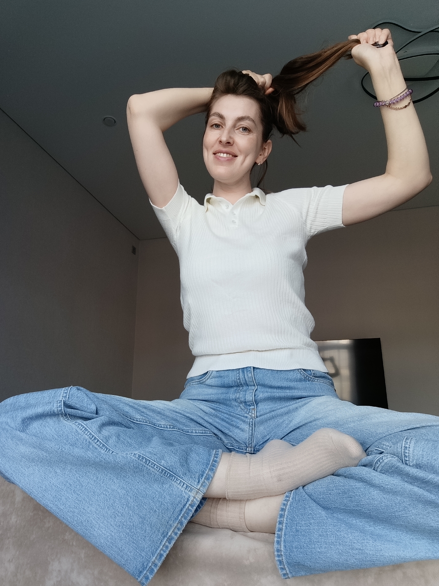 A woman is depicted in a medium shot, seated cross-legged on a light-colored upholstered surface, likely a bed or sofa, indoors. She is looking directly at the camera with a bright smile, showing visible dimples. Her brown hair is held up by both hands, as if she is in the process of tying it into a ponytail or bun.
She is wearing a cream or off-white ribbed short-sleeved polo shirt, light blue wide-leg jeans, and light beige ribbed socks. On her right wrist, she wears several bracelets, including a delicate gold chain and two purple beaded ones. Small, simple earrings are visible on her ears.
The background consists of plain, light gray walls and a matching ceiling, which features a small, circular recessed light fixture. A dark, rectangular object, possibly a television or monitor, is partially visible behind her to the right. The ambient lighting suggests it is daytime. The overall atmosphere conveys a sense of cheerfulness and relaxation. No text is visible in the image. The scene is set in Chaikovsky, Russia.