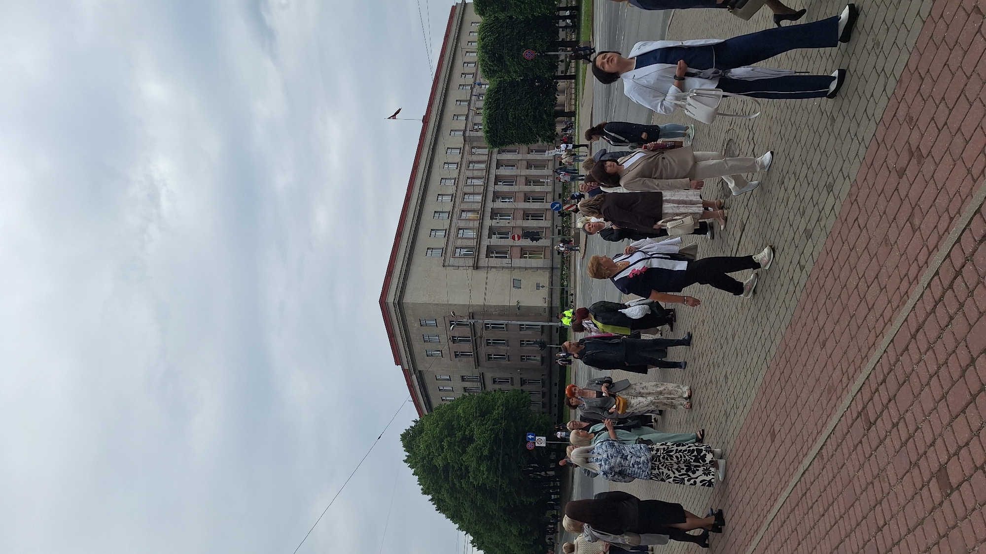 A group of people, primarily women dressed in casual to semi-formal attire, are gathered on a brick-paved sidewalk in front of a large, imposing, light-beige building with a red-tiled roof. The building exhibits a symmetrical facade with numerous windows arranged in horizontal rows across multiple stories. A Latvian flag flies from a pole atop the building.
The people are standing and walking, some in conversation, and appear to be observing something or waiting for an event. Several individuals are carrying bags or purses. Among the crowd, a person in a bright yellow vest is visible near the building entrance, possibly a security or event staff member. Lush green trees line the street in front of the building, and traffic lights and street signs are present in the background, indicating an urban setting. The sky is overcast and grey. The scene suggests a public gathering or a commemorative event taking place in a civic or administrative area of Riga, Latvia. The building's architecture hints at a governmental or institutional purpose.