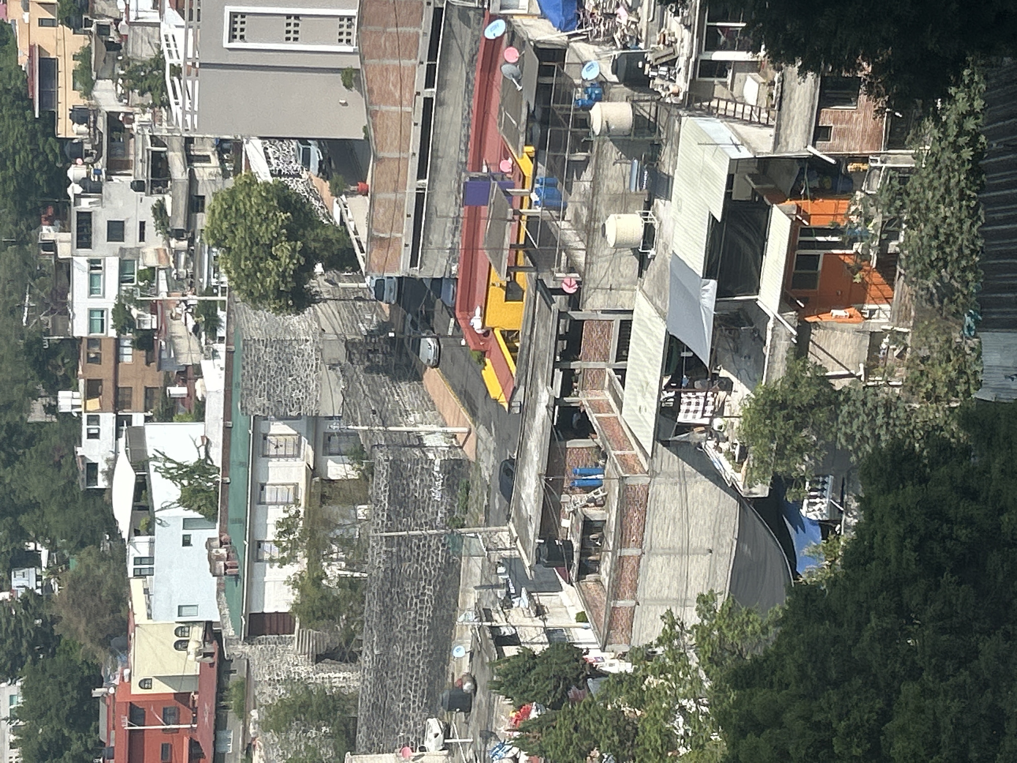 The image presents an aerial view of a dense residential area in Mexico City, Mexico. The scene is characterized by a mixture of building styles and materials, with structures closely packed together. Flat rooftops are visible, some adorned with satellite dishes and water tanks. The color palette is primarily muted, with occasional splashes of vibrant colors like red, yellow, and blue on some of the buildings. Trees and vegetation are interspersed throughout the urban landscape, providing a contrast to the concrete and brick structures. The overall impression is one of a densely populated, organically developed urban environment.