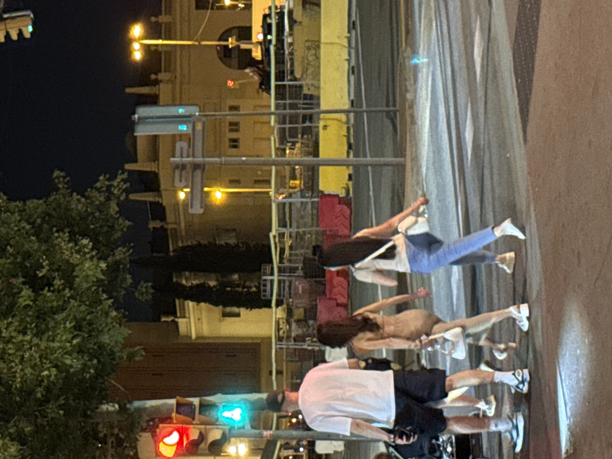 This image depicts a nighttime street scene in Barcelona, Spain. Several people are crossing a street at a pedestrian crossing, illuminated by streetlights and traffic signals. In the foreground, a man wearing a white t-shirt and dark shorts walks with a black camera bag. Behind him, two women are walking across the street. The woman on the left wears a light brown dress and carries a white handbag. The woman on the right wears a white t-shirt and blue jeans, also carrying a white handbag. Further in the background, traffic lights show a green signal, and building facades with illuminated windows are visible. Construction barriers, some with yellow and red elements, line the side of the street. The overall atmosphere suggests an urban environment at night with ongoing activity.