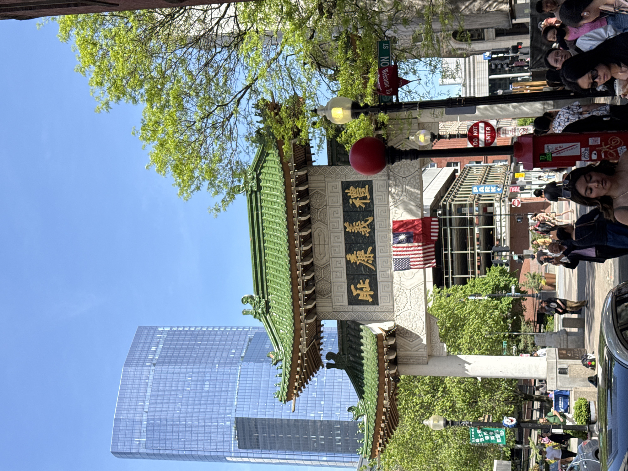The image captures a street scene in Boston's Chinatown, United States of America, featuring the iconic Chinatown Gate. The gate has a traditional green tiled roof and is adorned with gold Chinese characters. An American flag and the Taiwanese flag are visible behind the gate. A modern glass skyscraper towers in the background. A street sign for "Welcome To Washington St." is visible on a lamp post. People are walking in the background.