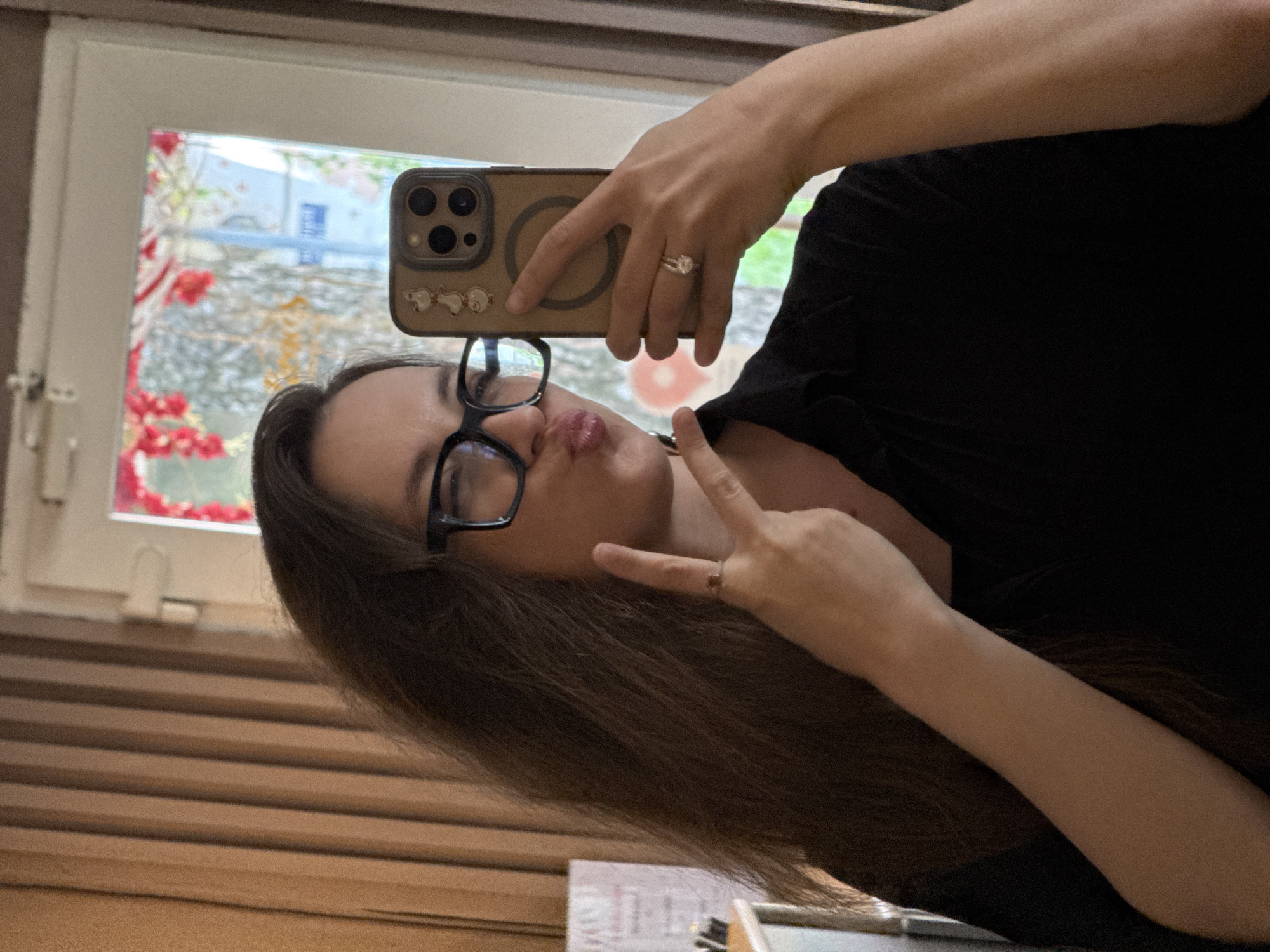 A woman with long, brown hair and glasses is taking a selfie in a room with beige curtains and a doorway reflecting a street scene. She is wearing a black shirt and making a peace sign with her right hand. Her left hand is holding a smartphone. She is also wearing two rings on her left hand. The background shows a window with red and white flowers.