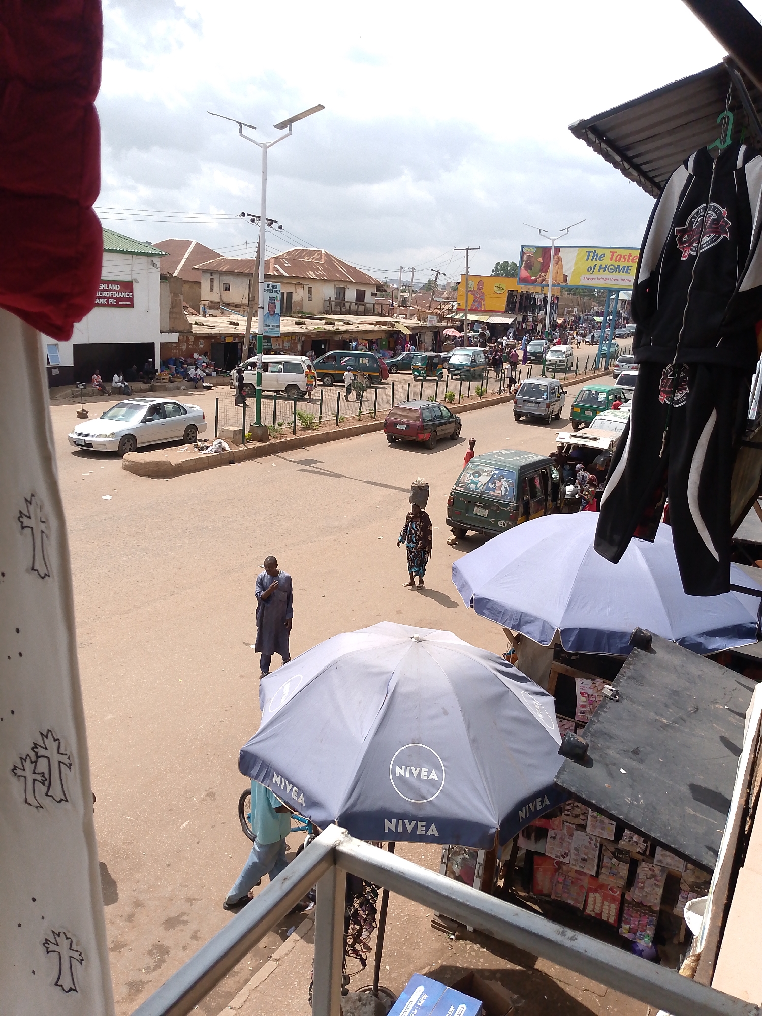 This media file presents a bustling street scene in Bukuru, Nigeria, viewed from an elevated perspective, possibly a balcony. The foreground includes a metal railing and a white fabric with embroidered cross motifs, partially obscuring the view.
The central feature is a wide, busy street filled with a mix of pedestrians and vehicles. Several cars, including a white sedan, various green minibuses, and a tricycle (keke napep), are seen driving or parked. Numerous people are walking along the road and sidewalks. Notable figures include a woman expertly balancing a large load on her head and a man in a long, dark tunic standing in the middle of the street.
Along the left side of the street, a white building houses "HIGHLAND MICROFINANCE BANK Plc," and several individuals are gathered in a shaded area beneath it. To the right, three large market umbrellas are visible, two prominently featuring the "NIVEA" brand logo, indicating commercial activity. A black and white jacket and trousers hang on a display rack in the immediate right foreground, likely merchandise for sale.
In the mid-ground and background, more buildings line the street, some with traditional reddish roofs and others appearing to be shop fronts. Large billboards are prominent, displaying text such as "The Taste of HOME Always brings them home" and "ONGA," suggesting food or consumer product advertisements. A sign for a "Mall" is also visible. Modern solar-powered streetlights are installed along the roadside.
The sky is overcast, indicating a cloudy daytime, but the overall illumination is bright. The scene captures the lively, everyday activity of an urban or semi-urban commercial area in Nigeria.