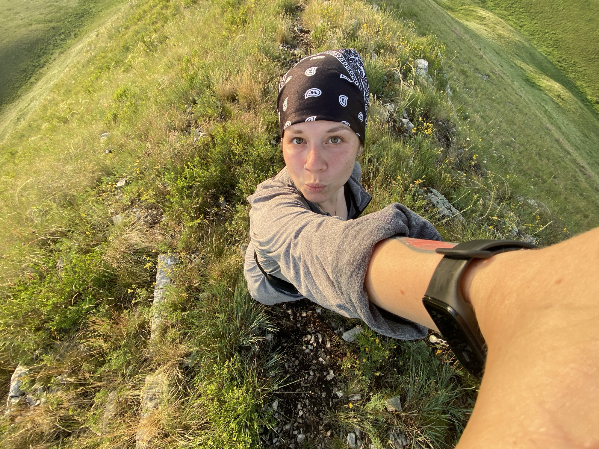 A woman in a grey hoodie and a black bandana on her head is taking a selfie on a grassy hillside. She is wearing a black fitness tracker on her left wrist. The background is a sloping green hill with patches of rocks and yellow wildflowers. The scene is outdoors in daylight, with the sun casting a warm glow on the landscape. The location is in Novogafarovo, Russia.
