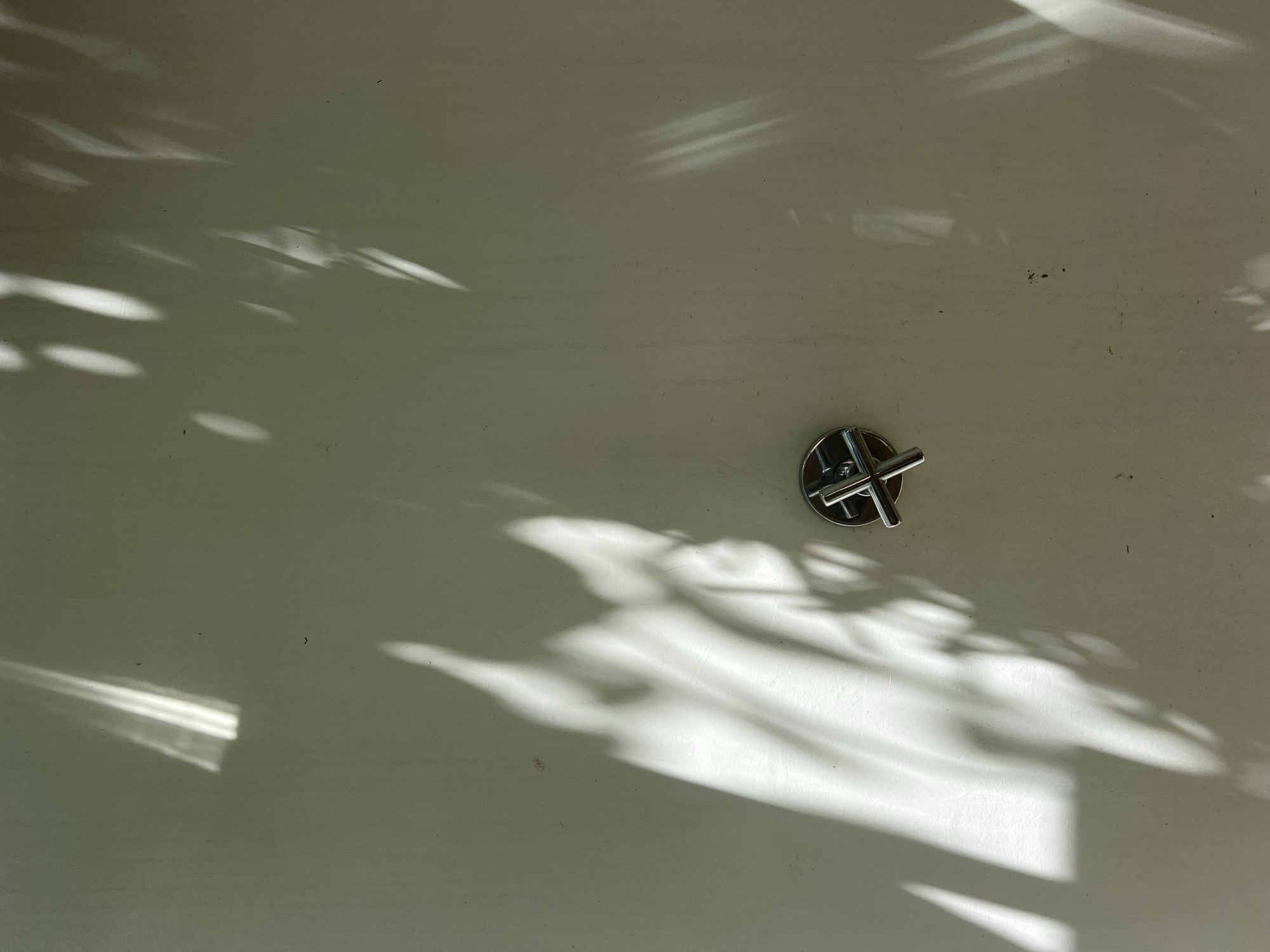 The image captures a close-up, high-angle view of a light-colored, possibly off-white or very light grey, planar surface. This surface could be a wall, a tabletop, or a deck, and it shows minor imperfections, including small dark specks or dirt.
A significant portion of the scene is dominated by the intricate interplay of natural sunlight and shadows. Distinct, organic shadow patterns, resembling those cast by tree leaves or branches, stretch across the surface, creating a dynamic visual contrast between illuminated areas and shaded regions. Some parts of the shadows are soft and diffused, while others show sharper edges, indicating direct sunlight.
Towards the right-center of the frame, a small, metallic object is visible. It appears to be a polished, silver-colored knob or fixture with a distinctive cross-like or plus-sign shape on its top. This object also casts a small, crisp shadow on the surface directly beneath it.
The strong, well-defined shadows and bright highlights indicate a sunny time of day. No specific activity or people are present, making the image a study of light, shadow, and texture on a utilitarian surface. There is no visible text in the image. The overall impression is one of calm observation, possibly in an outdoor or naturally lit indoor setting where foliage is nearby.