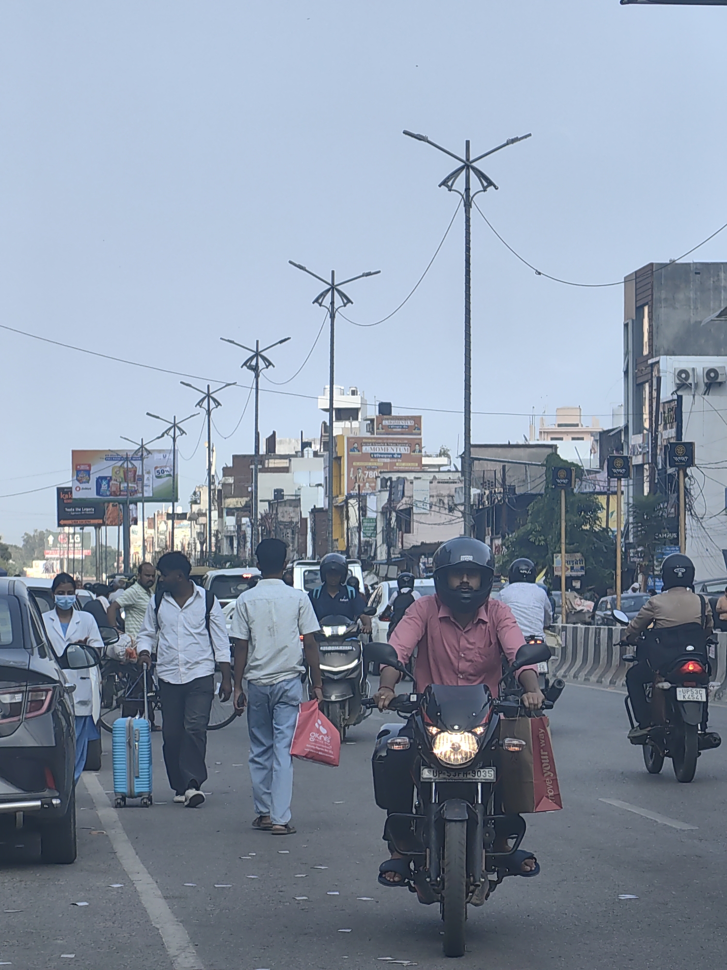 The image captures a busy street scene in Gorakhpur, India, likely during the daytime given the illumination. In the foreground, a person wearing a pink shirt and a black helmet is riding a motorcycle. The motorcycle has its headlights on and a red shopping bag with white text that reads "move your way" is attached to the front. Behind this motorcycle, another person on a motorcycle is visible, also wearing a helmet and a light brown jacket, with the license plate "UP53C K4541" clearly visible. Further back in the traffic, a person in a white shirt and dark pants is walking with a blue suitcase on wheels. To the left, a person in a white coat and blue scrubs, possibly a healthcare worker, is standing near a dark-colored car. The street is lined with buildings and billboards. Several tall streetlights with multiple lamps are also visible, spaced along the road. The overall impression is one of congested urban traffic.