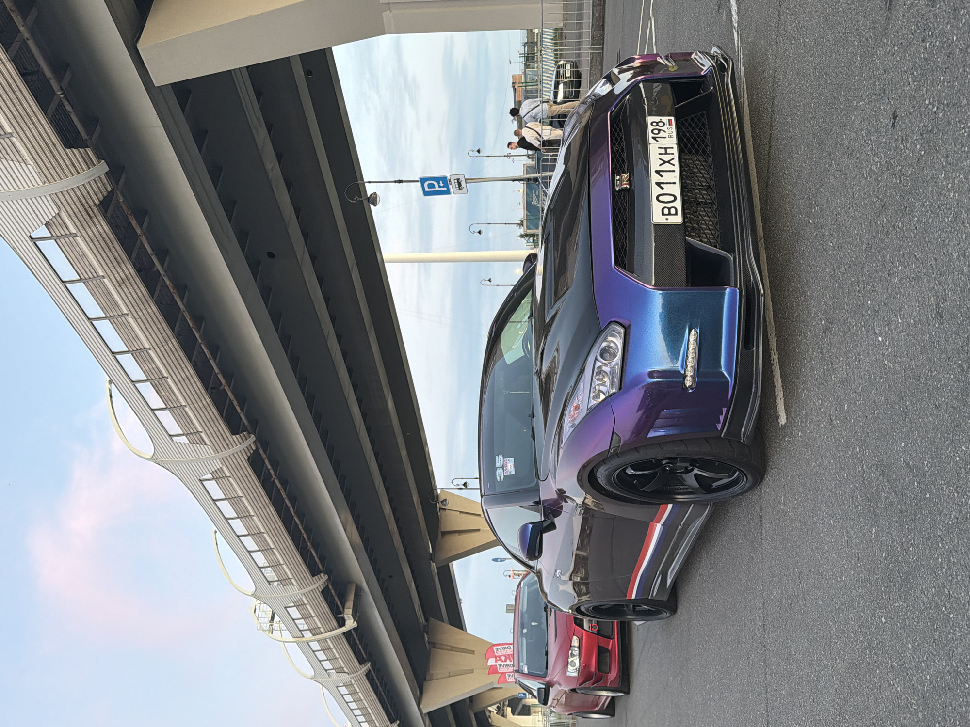 A highly modified, iridescent purple and blue Nissan GT-R is parked on asphalt, partially under a concrete overpass. The car has a black hood, black wheels, and a red, white, and blue stripe along its side. A red Mitsubishi Lancer Evolution is parked behind it. Several people are visible in the background near a street sign that indicates parking and bus access. The sky is partly cloudy.