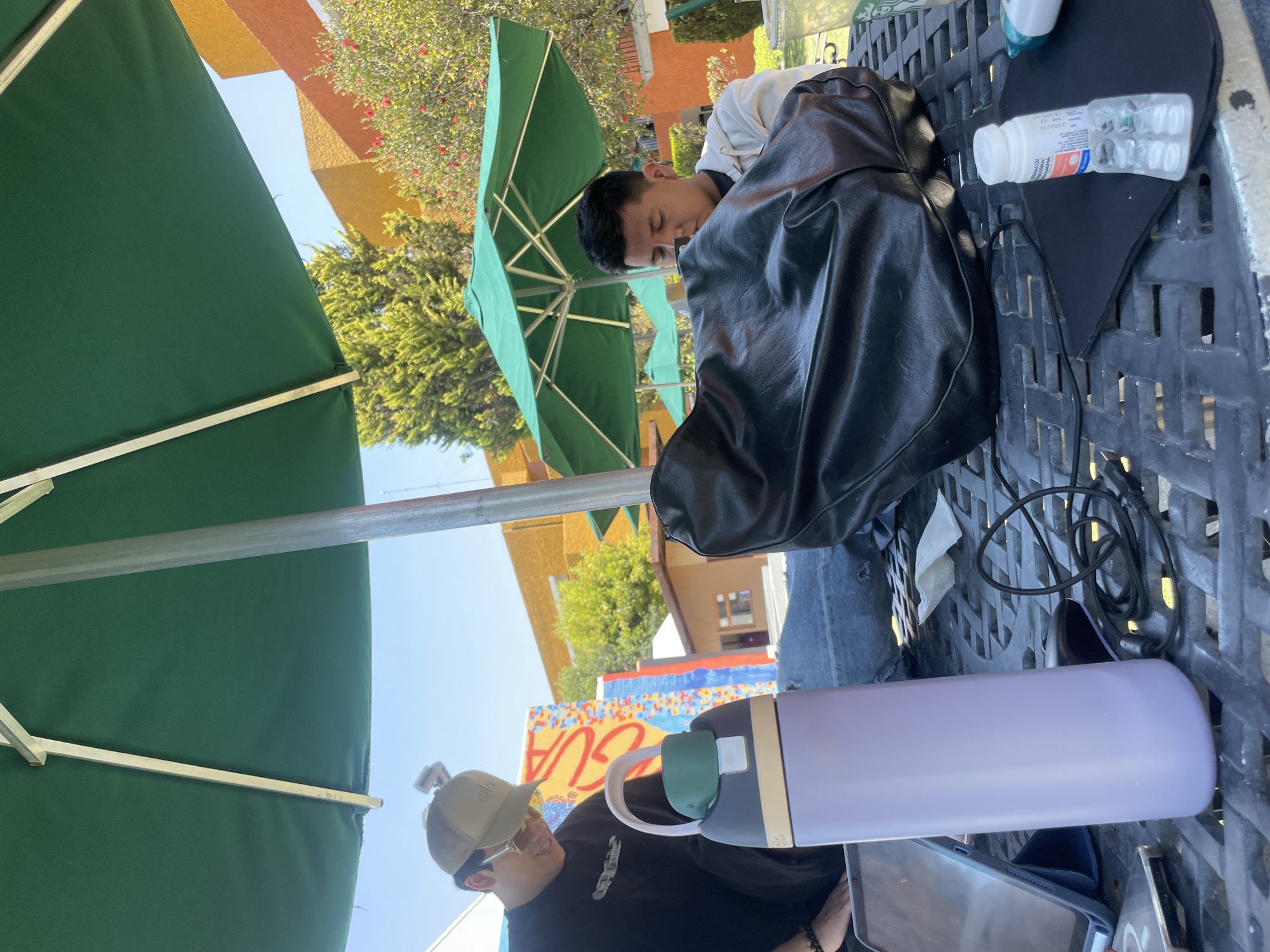 An outdoor scene in San Andrés Cholula, Mexico, depicts two males seated at a dark-colored, textured patio table under a large green umbrella on a bright, sunny day.
To the left, one male wears a beige baseball cap with a camera mounted on top, yellow-tinted sunglasses, and a black long-sleeved shirt with "OFF-WHITE" text. He has a black bracelet on his left wrist and is looking down towards the table. Adjacent to him on the table is a light purple insulated water bottle with a green mouthpiece and beige handle, a tablet in a blue case displaying a screen, and a partially visible smartphone.
To the right, a second male, with short dark hair, wears a light-colored hooded sweatshirt and is looking down, partially obscured by a large black soft-sided bag resting on the table in front of him.
Between the two individuals on the table are tangled black cables. Further to the right, on a black mat, are a small white cylindrical bottle with "FARMACIA" visible on its label and an empty blister pack of pills. A clear plastic cup with a green logo, containing a light-colored beverage, and a crumpled white napkin are also present.
In the background, a building with a vibrant mural featuring red, yellow, blue, and white elements, including the word "AGUA," is visible. Another building with warm orange and yellow walls stands further back, alongside green trees and shrubs with red blooms. Several other green patio umbrellas are visible in the mid-ground.