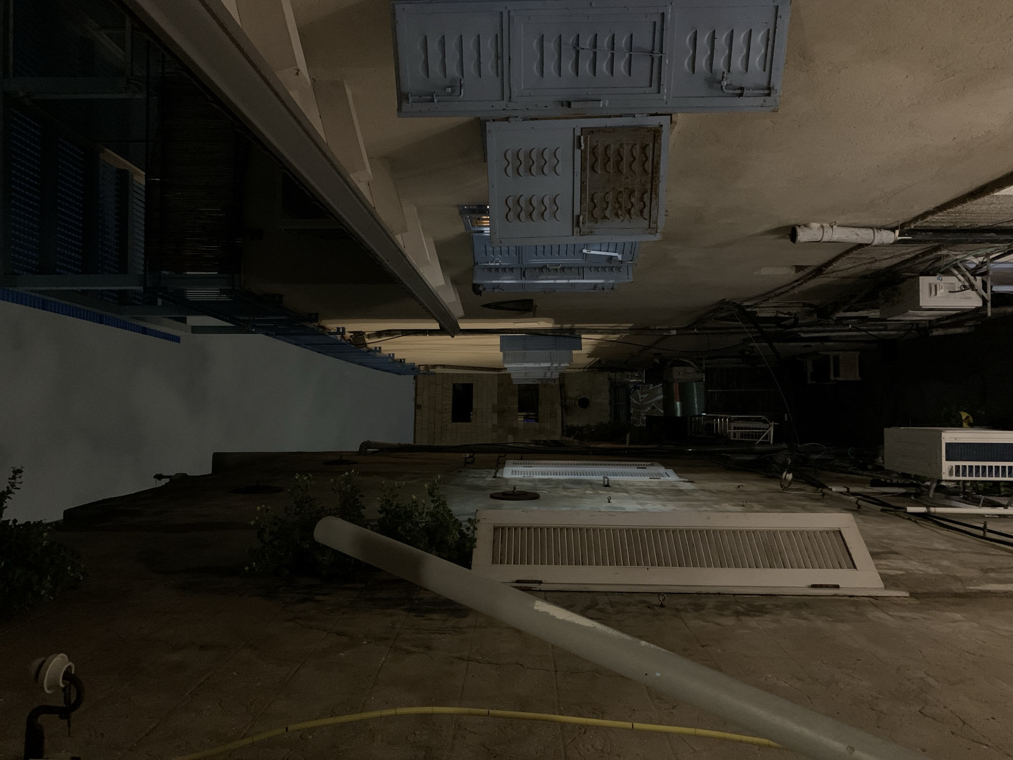 This image depicts a narrow urban alleyway at night in Tel-Aviv, Israel, viewed from a slightly elevated perspective looking down its length. The scene is devoid of individuals.
On the left, a textured, light brown or beige building wall rises steeply. A thick, off-white drainpipe runs diagonally downwards across this wall, originating from the upper left. Below this pipe, a rectangular window with a vertical white louvered shutter is visible, angled slightly outwards. Above the shutter, green foliage protrudes from the wall. A yellow flexible conduit runs vertically along the far upper left. Near the bottom, an outdoor air conditioning unit is mounted on the wall.
On the right, a lighter beige or cream-colored building wall runs parallel to the left wall. Multiple windows are present, each fitted with light blue louvered shutters. Several shutters are partially open, revealing dark interiors. One shutter unit features two rows of small, horizontal, wavy ventilation slits. Above the windows, a dark grey gutter runs horizontally along the roofline. Above the gutter, a dark, open-sided structure with a slatted roof is visible, suggesting a balcony or pergola area on a higher floor. Near the bottom of this wall, another outdoor air conditioning unit is mounted.
The alley floor is not visible, obscured by the close proximity of the walls. Numerous dark cables and smaller pipes are affixed to both walls, particularly noticeable in the mid-ground. The alley recedes into deep shadow and appears to narrow in the background, where faint outlines of additional, darker buildings are discernable under a dark, overcast night sky. Artificial light illuminates portions of the building walls, contrasting with the dark sky and deeper shadows within the alley.