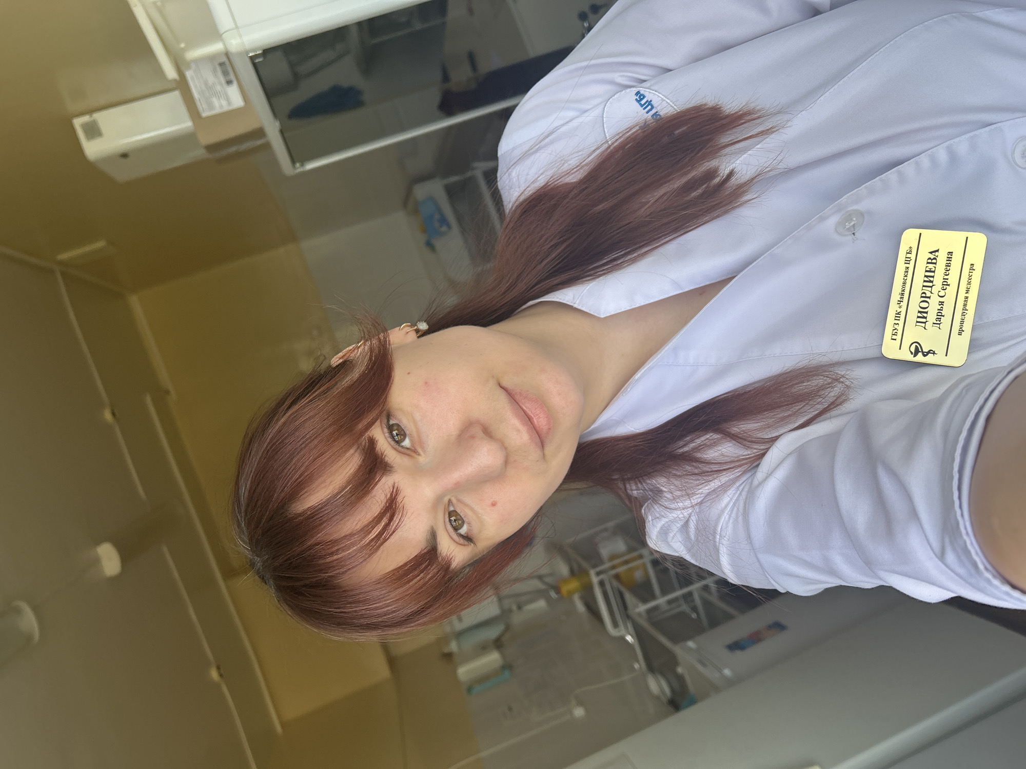 A young woman with reddish-brown hair and bangs is wearing a white lab coat and looking at the camera. A name tag on her coat reads "ГБУЗ ПК 'Чайковская ЦГБ' ДИОРДИЕВА Дарья Сергеевна процедурная медсестра". The location on the tag indicates she is in the city of Чайковский (Chaykovsky) in the Perm Krai region of Russia, not Фоки. The background suggests a medical setting, possibly a hospital or clinic, with medical equipment and cabinets visible. The image is slightly tilted.