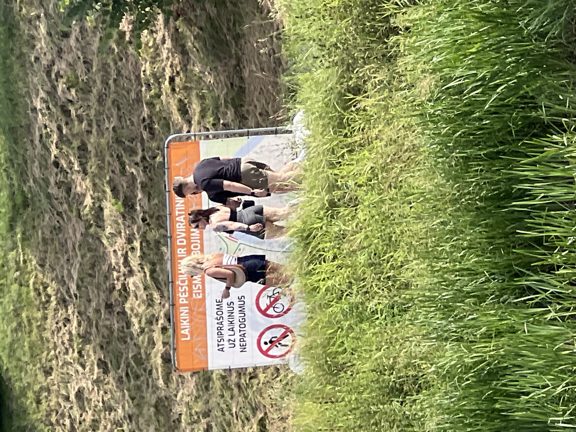 Three individuals are standing in front of an orange and white sign in a grassy area. The sign has Lithuanian text and two circular prohibition symbols, one depicting a walking person and the other a bicycle. The individual on the left is a woman with blonde hair wearing a striped top and denim shorts, carrying a tan shoulder bag. The person in the middle is a woman with dark hair wearing a black top and shorts, with visible tattoos on her arm and leg. The person on the right is a man wearing a black t-shirt and khaki shorts. The background consists of a grassy slope with some dry vegetation and trees. The scene appears to be outdoors in a natural setting. The text on the sign, when combined with the context of the prohibition symbols, suggests it is a notice regarding restrictions on pedestrian and bicycle access. The location is the Vilnelė River trail, specifically near the bridge leading to the Paupys district of Vilnius, Lithuania.