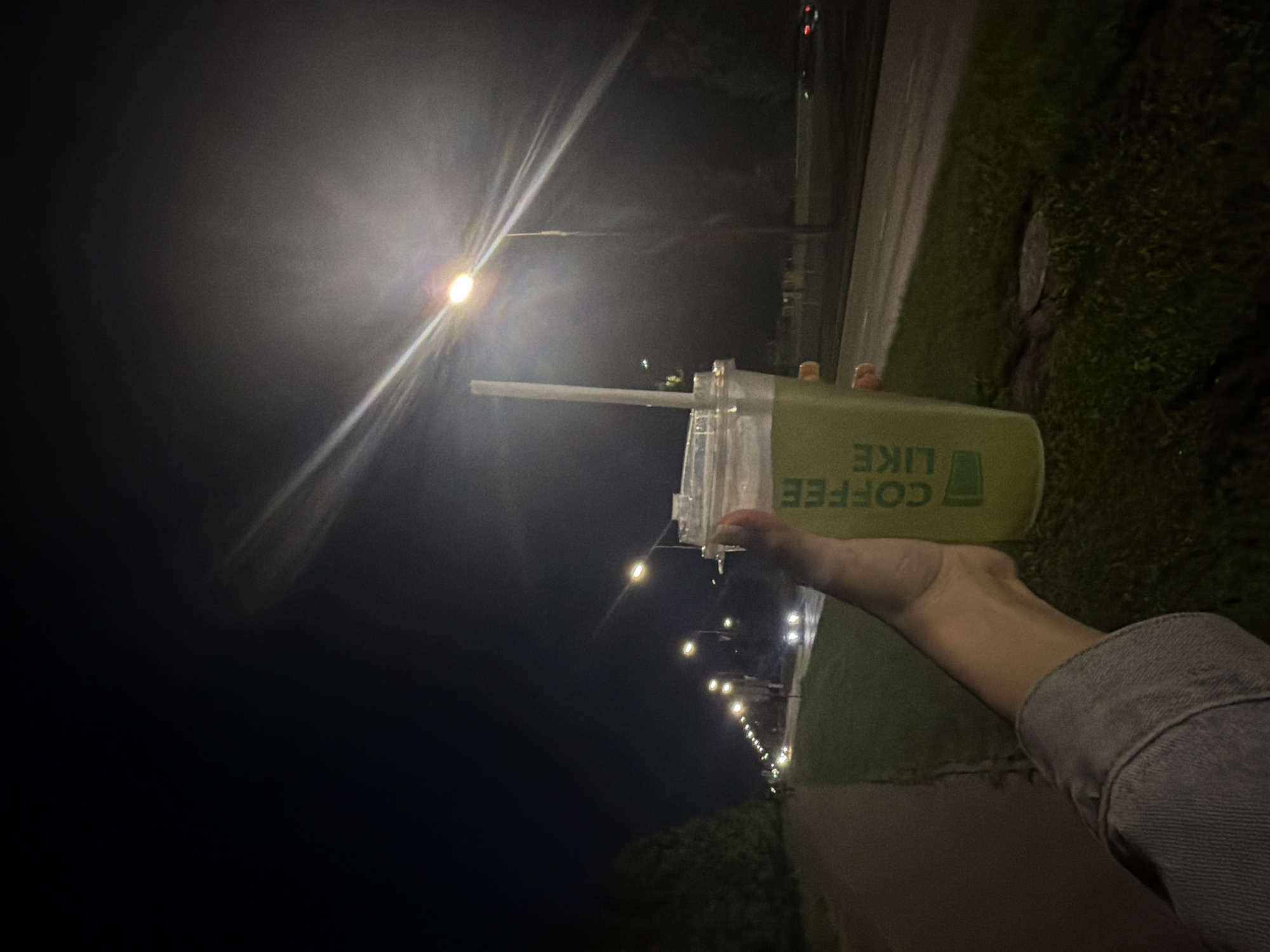 The image shows a person's hand holding a translucent plastic cup with a green beverage and a straw, in front of a street scene at night. The cup has "COFFEE LIKE" written on it in green lettering. The background features a road with several streetlights and passing cars. A sidewalk or path is visible on the left side of the image, and trees line the edge of the road. The sky is dark. The presence of streetlights and the arrangement of the road and sidewalk suggest an urban or suburban environment. The overall scene conveys a sense of a night-time excursion, possibly a walk or commute. The location is identified as Naberezhnye Chelny, Russia.