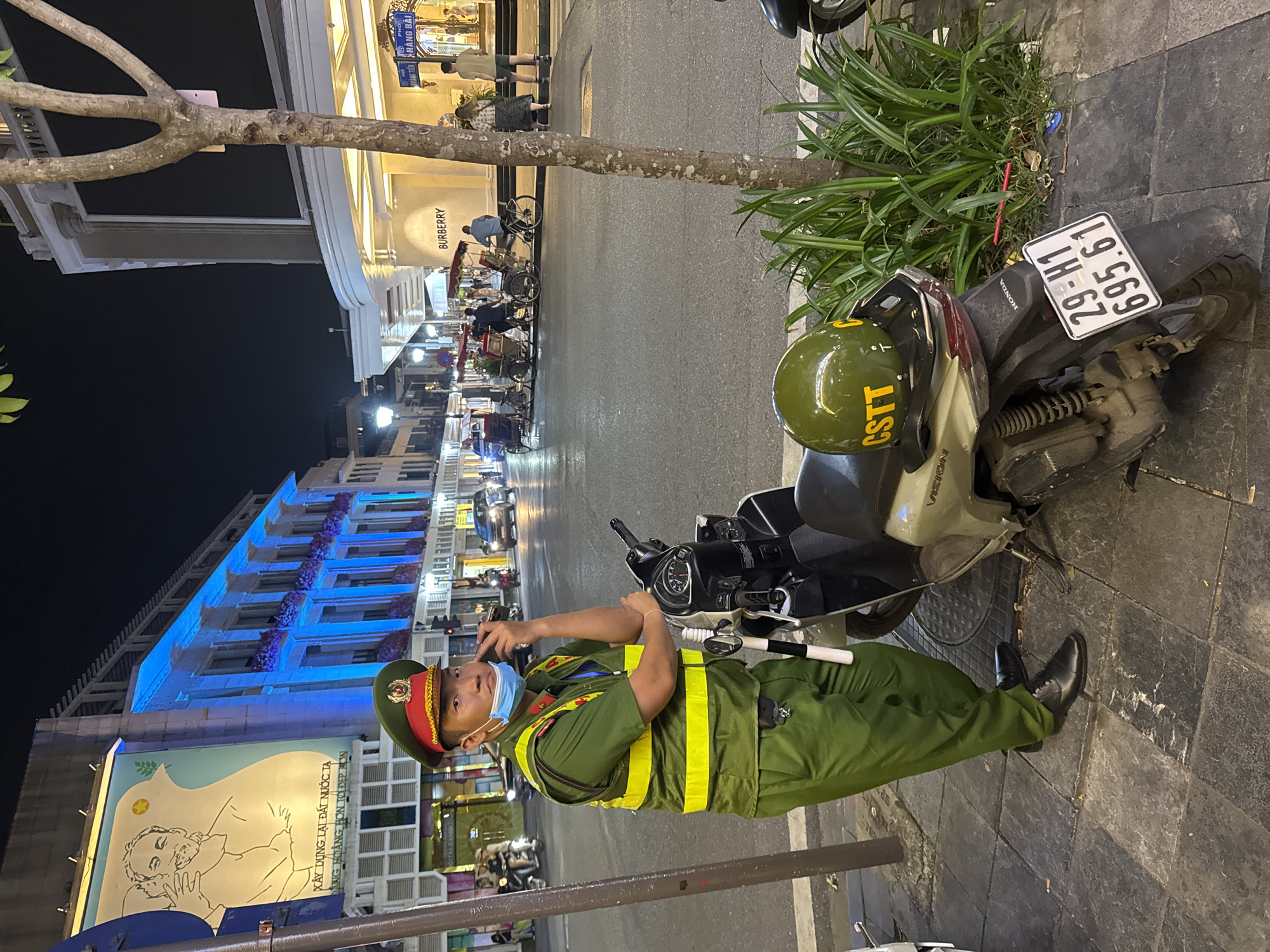 This media file captures a night scene on a city street, likely in Hanoi, Vietnam, as suggested by the text "XÂY DỰNG LẠI ĐẤT NƯỚC TẠ" and the presence of a uniformed individual that appears to be a traffic or public order officer. The officer is standing next to a motorcycle, wearing a green uniform with a yellow reflective vest, a peaked cap, and a face mask. They are holding a phone to their ear, seemingly engaged in a conversation.
The background reveals a brightly lit building with blue accents and balconies with purple flowers, suggesting a commercial or residential area. Further down the street, other buildings and shops are visible, including a store with "BURBERRY" signage. Several pedicabs and other motorcycles are parked or moving in the background, indicating a bustling urban environment. A large mural depicting a stylized portrait of a man is also visible on a building.
The lighting suggests it is nighttime, with artificial lights illuminating the street and buildings. The ground is paved with dark tiles. The overall atmosphere appears to be that of a typical evening in a Vietnamese city, with the officer on duty, maintaining order amidst the urban activity. The motorcycle has a license plate that reads "29-H1 695.61".