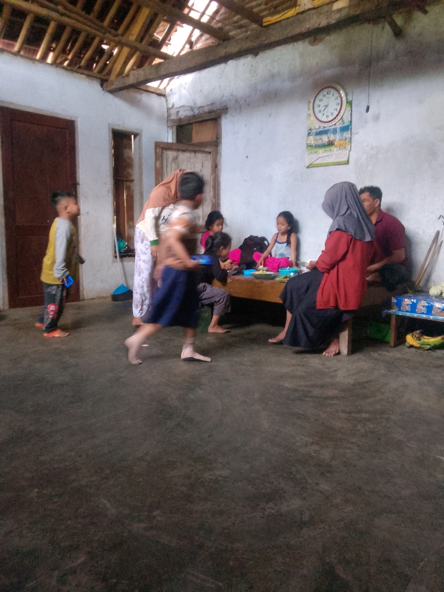 The image depicts an indoor scene, likely a modest dwelling, in Krucil, Indonesia. Several individuals, including children and adults, are present. The environment appears somewhat sparse, with a concrete floor and whitewashed walls. A wooden beam structure is visible along the ceiling, suggesting a traditional construction.
In the foreground, a young boy wearing a yellow and grey long-sleeved shirt and patterned pants is standing, holding a blue object. To his right, a blurred figure of a child in a dark blue skirt and a striped shirt is running.
Further back, a woman in a light-colored garment and a long patterned skirt stands near a table. On the table, several children are gathered. One girl wears a light blue top and dark pants, another, with dark hair, is partially visible. A woman wearing a grey headscarf and a red top is seated at the table, facing away from the camera, and a man in a maroon shirt is seated next to her.
The table itself is laden with various items, including plates of food and what appear to be bowls. To the right of the table, a small blue stool or table holds what looks like a packaged item and bananas. A clock is mounted on the wall, displaying the time. Two doorways are visible in the background, one a darker, solid door and another a more open, framed entrance. A broom with a blue handle leans against the wall.
The overall impression is of a communal gathering or meal in a simple, rural Indonesian home. The lighting is dim, suggesting it might be evening or a cloudy day.
