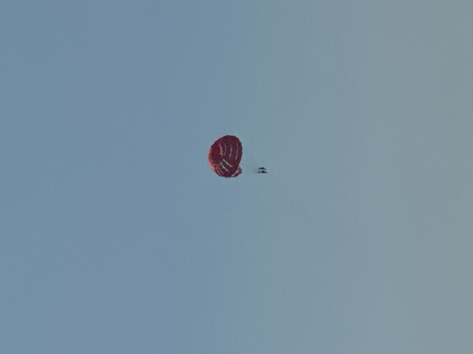 A single person is parasailing high in a clear, light blue sky. The parasail is a vibrant red with white stripes and is inflated, holding the person aloft. The person is suspended beneath the parasail by a harness and lines. The scene appears to be taking place over the sea, as is typical for parasailing activities in tourist destinations like Budva, Montenegro. The vast expanse of the sky dominates the image.