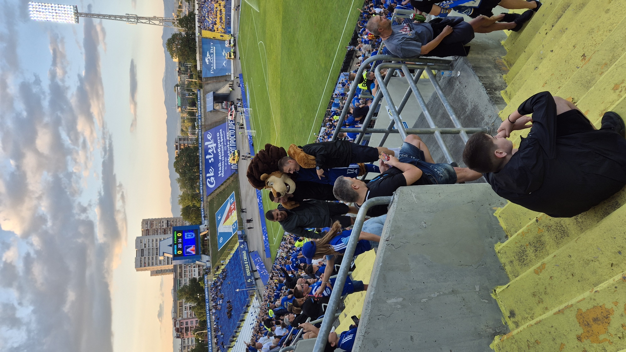 The image depicts a scene at a football stadium in Sofia, Bulgaria, likely during a match. In the foreground, a group of spectators are seated and standing on yellow-painted concrete steps, with metal railings separating the seating sections. A young person in a black hoodie is crouched on the steps, looking to the side.
Further up the steps, a mascot dressed as a lion is interacting with several individuals, including two men in dark jackets and another man in a dark t-shirt. The mascot has its arm around one of the men. Other spectators, many wearing blue, are visible in the background, filling the stands.
The stadium features a green football pitch, a scoreboard displaying "0 20 16 0," and several buildings and a tall stadium light visible in the distance. Advertising banners are present along the upper sections of the stadium. The sky above is partly cloudy with a warm, late afternoon light. The overall atmosphere suggests a sporting event is underway or has recently concluded.