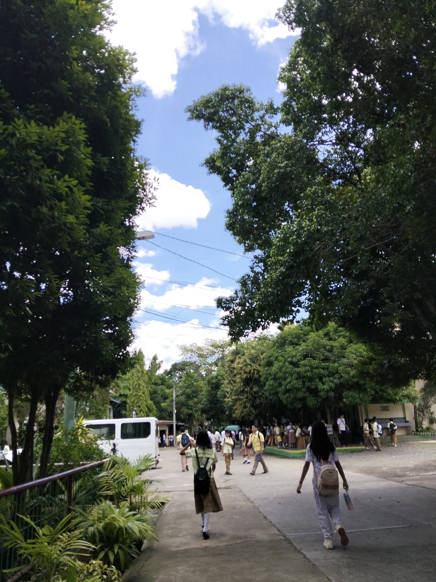 The image depicts a bright, sunny day with a clear blue sky dotted with fluffy white clouds. The scene is set outdoors, likely in a school or university campus, given the presence of students in uniform walking along a paved pathway. Lush green trees flank the pathway, providing shade and a natural, verdant environment. In the background, more students can be seen gathered, suggesting a school day in progress.
The primary subjects are the students, most of whom are walking away from the camera. One student in the foreground on the right is wearing a white shirt and light-colored pants, carrying a backpack and a water bottle. Another student in the middle of the pathway is wearing a green and white striped shirt with a brown skirt and a backpack. Students in the background are in various uniforms, some in yellow shirts and brown pants, and others in white tops and brown skirts, typical school attire in the Philippines.
An object of note is a white van parked on the left side of the pathway, possibly a school service vehicle. Other details include a lamppost and power lines overhead, common in urban and suburban settings. The overall atmosphere seems peaceful and ordinary, indicative of a normal school day. The vibrant greenery and bright sky suggest it's daytime, likely mid-morning or early afternoon, with pleasant weather. While no specific text is clearly legible, the general setting points to an educational institution in Davao City, Philippines, due to the context provided.