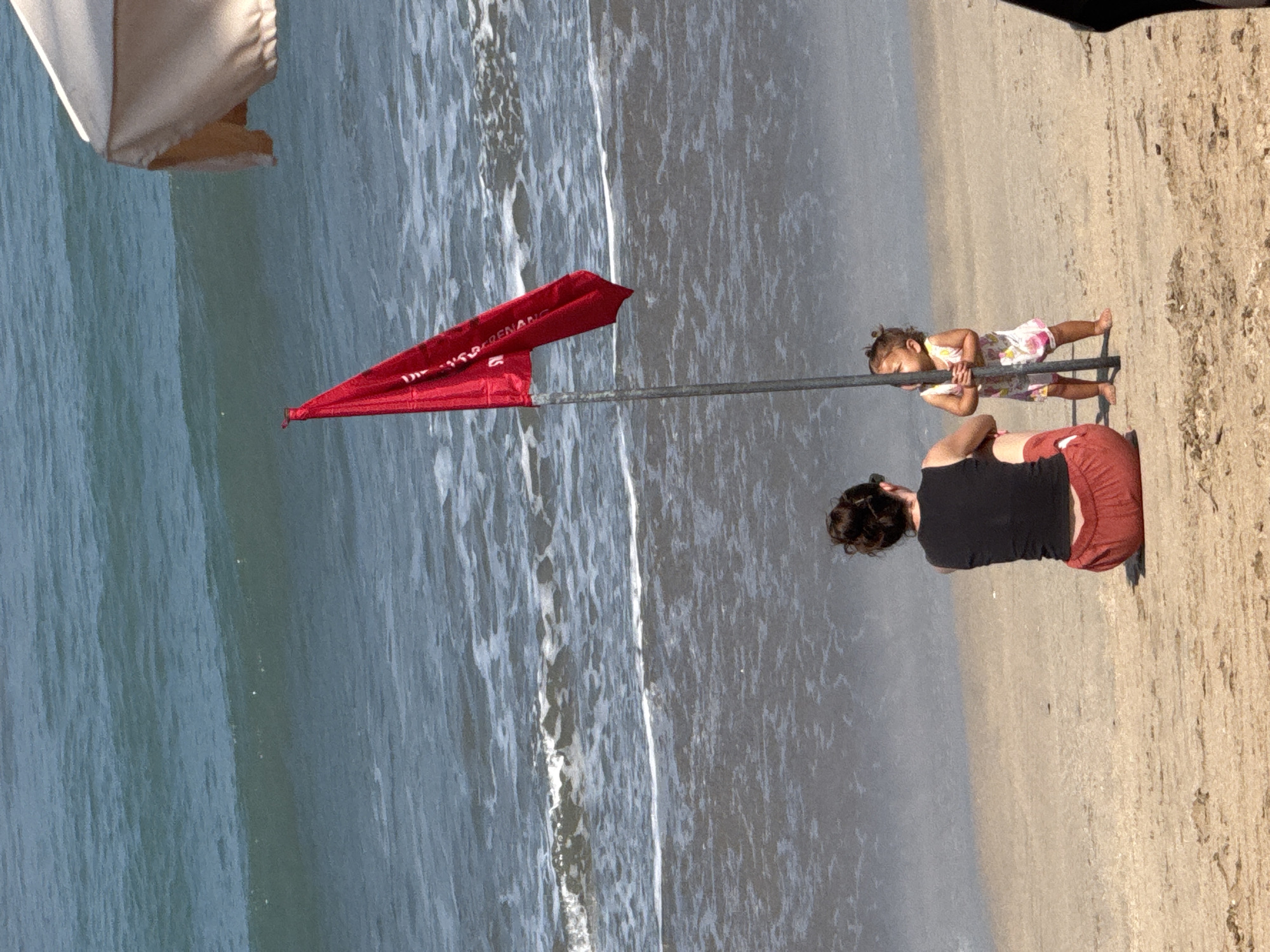 A rear view of a woman and a toddler are depicted sitting and standing on a sandy beach with the ocean in the background. The woman is seated, facing away from the camera, wearing a black sleeveless top and red shorts. The toddler is standing to her right, also facing away from the camera, wearing a light-colored dress with floral patterns. They are positioned near a metal pole with a red flag attached to it, which is partially obscured by the toddler. The ocean exhibits moderate waves and a foamy shoreline. A portion of a beige beach umbrella is visible in the upper right corner of the frame. The scene suggests a sunny day at a beach in Kuta, Indonesia, potentially a public beach with safety warnings indicated by the red flag.